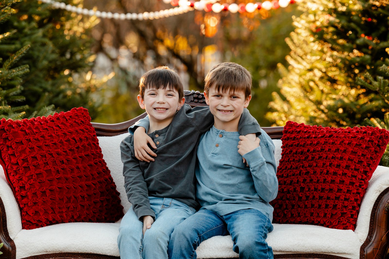 Two young boys sitting close together on a decorated outdoor sofa, smiling, with red pillows on either side, during a sunset in an outdoor winter or holiday setting.