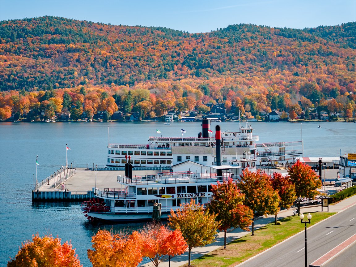 Ferry boat docked at a pier along a lake, surrounded by fall foliage, with a mountain in the background and a walkway lined with colorful trees in the foreground.