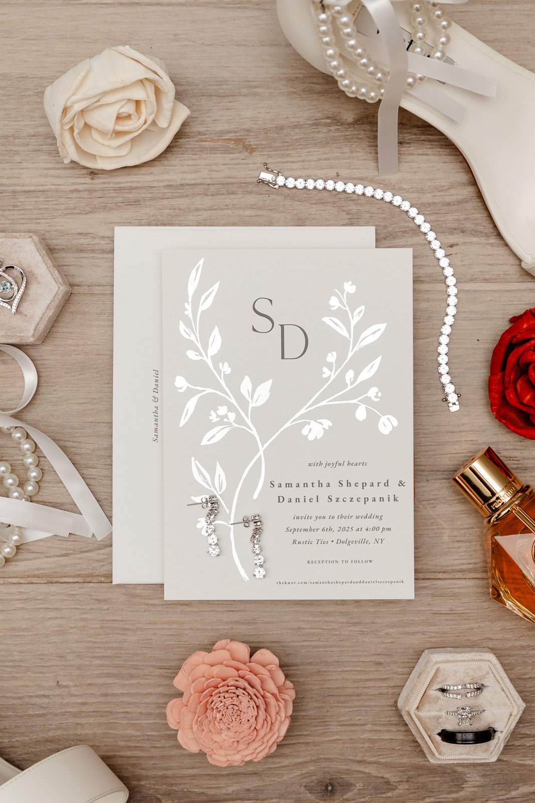 Wedding invitation surrounded by jewelry, flowers, perfume, and accessories on a wooden surface.