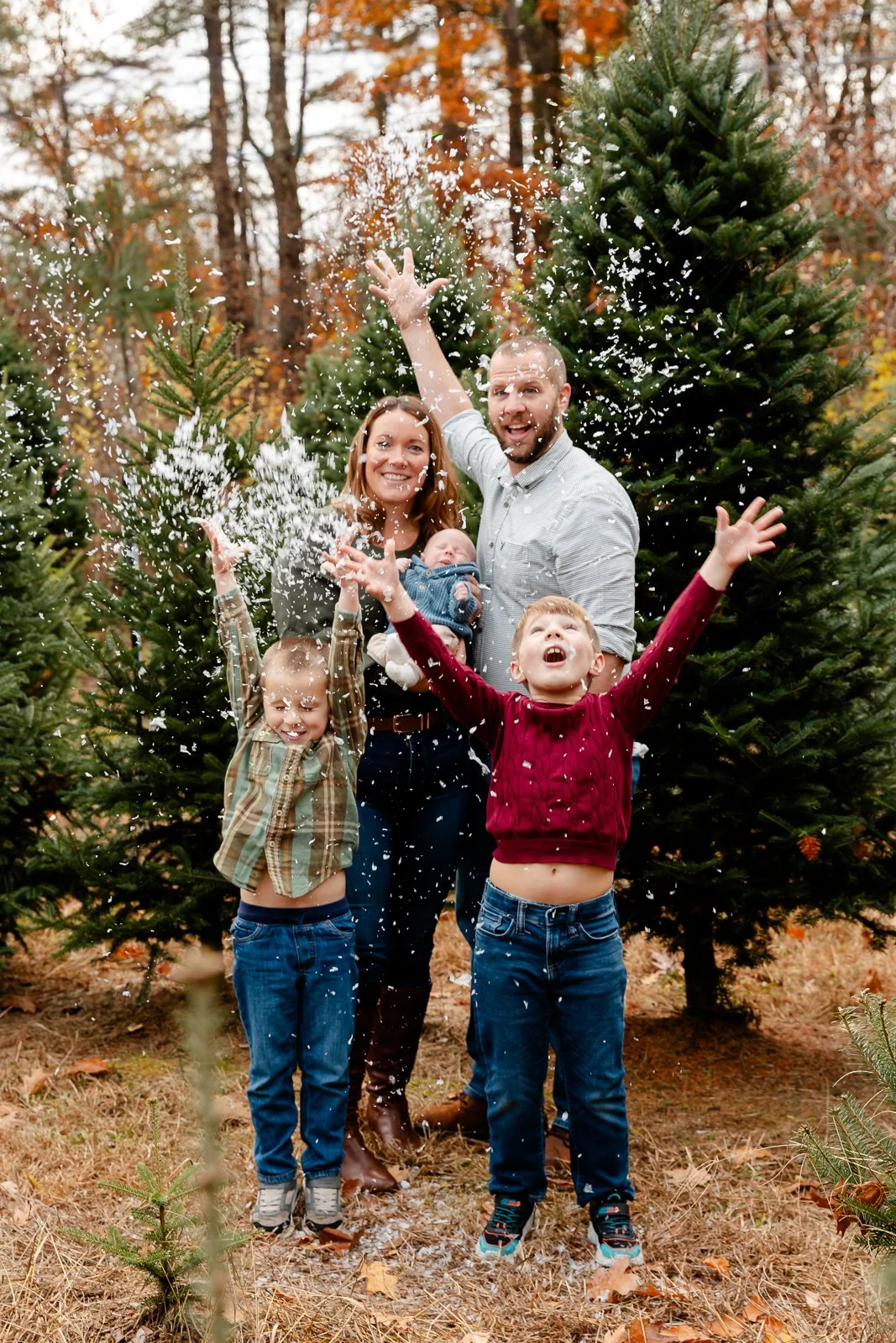 Family celebrating outdoors among Christmas trees, throwing snow or artificial snow in the air, smiling and laughing, autumn leaves on the ground.