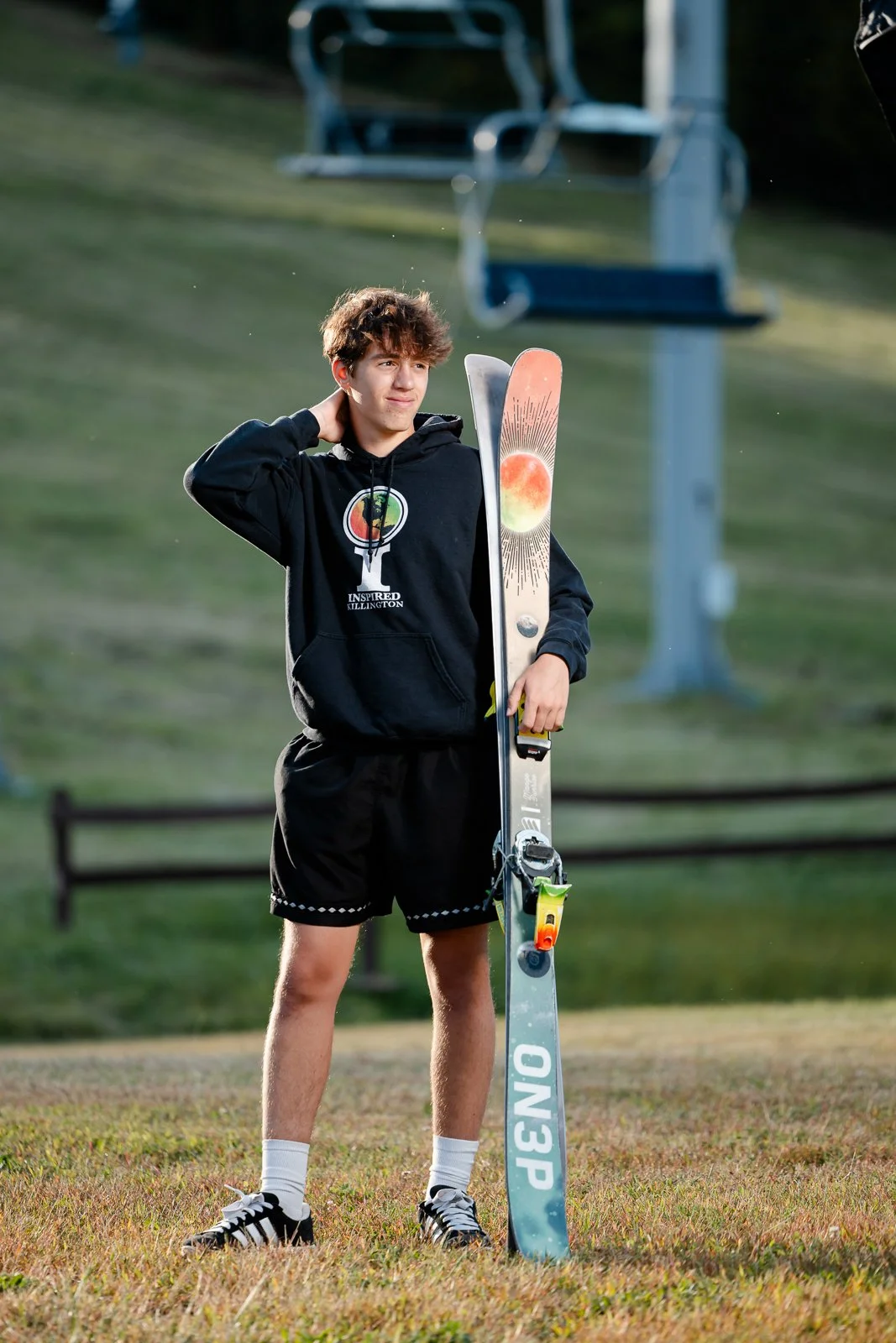 A teenage boy standing on grass holding a pair of skis vertically, with one hand resting on his neck. He is wearing a black hoodie, black shorts, white socks, and black sneakers. In the background, there are blurred green hills and ski lift chairs.