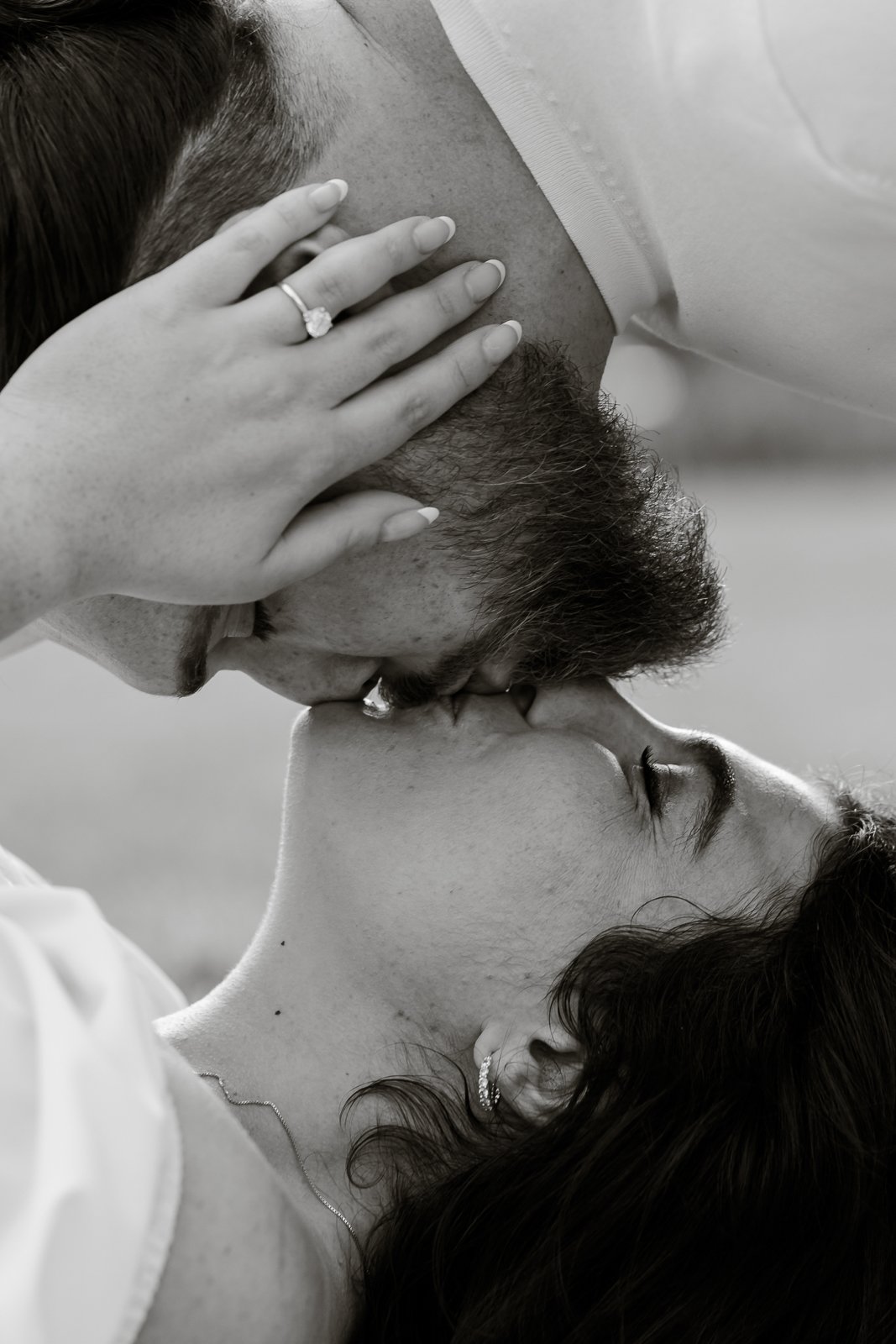 A black and white photo of a woman and a man sharing a kiss. The woman has her hand on the man's head, showing an engagement ring on her finger. The man has a beard, and the woman has earrings and a necklace.