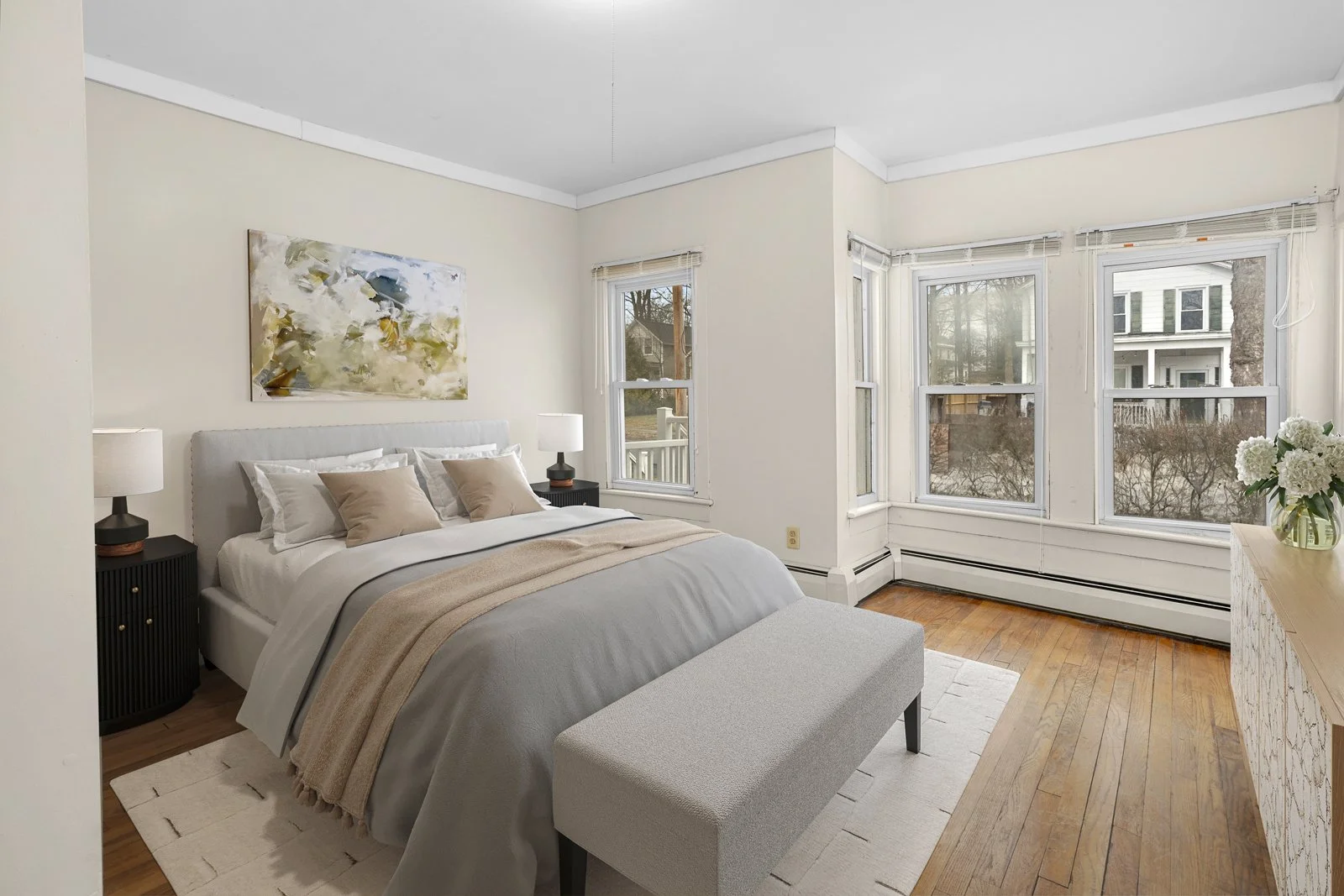 A bedroom with a white bed, beige and gray bedding, two black nightstands with white lamps, a light-colored bench at the foot of the bed, hardwood floors, and large windows with views of neighboring houses and trees.