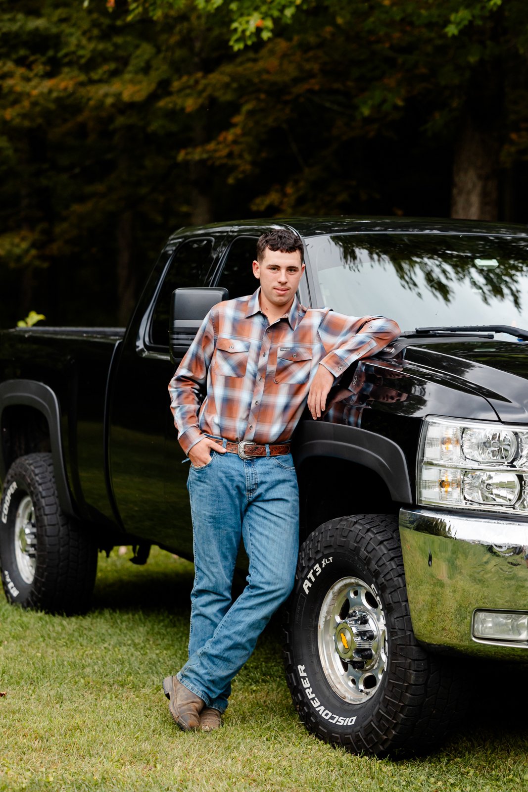 A young man in a plaid shirt and blue jeans leaning against a black pickup truck, outdoors in a grassy area with trees in the background.