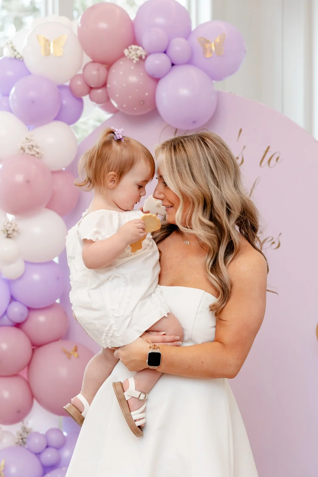 A woman holding a young girl during a celebration with pink and purple balloons in the background.