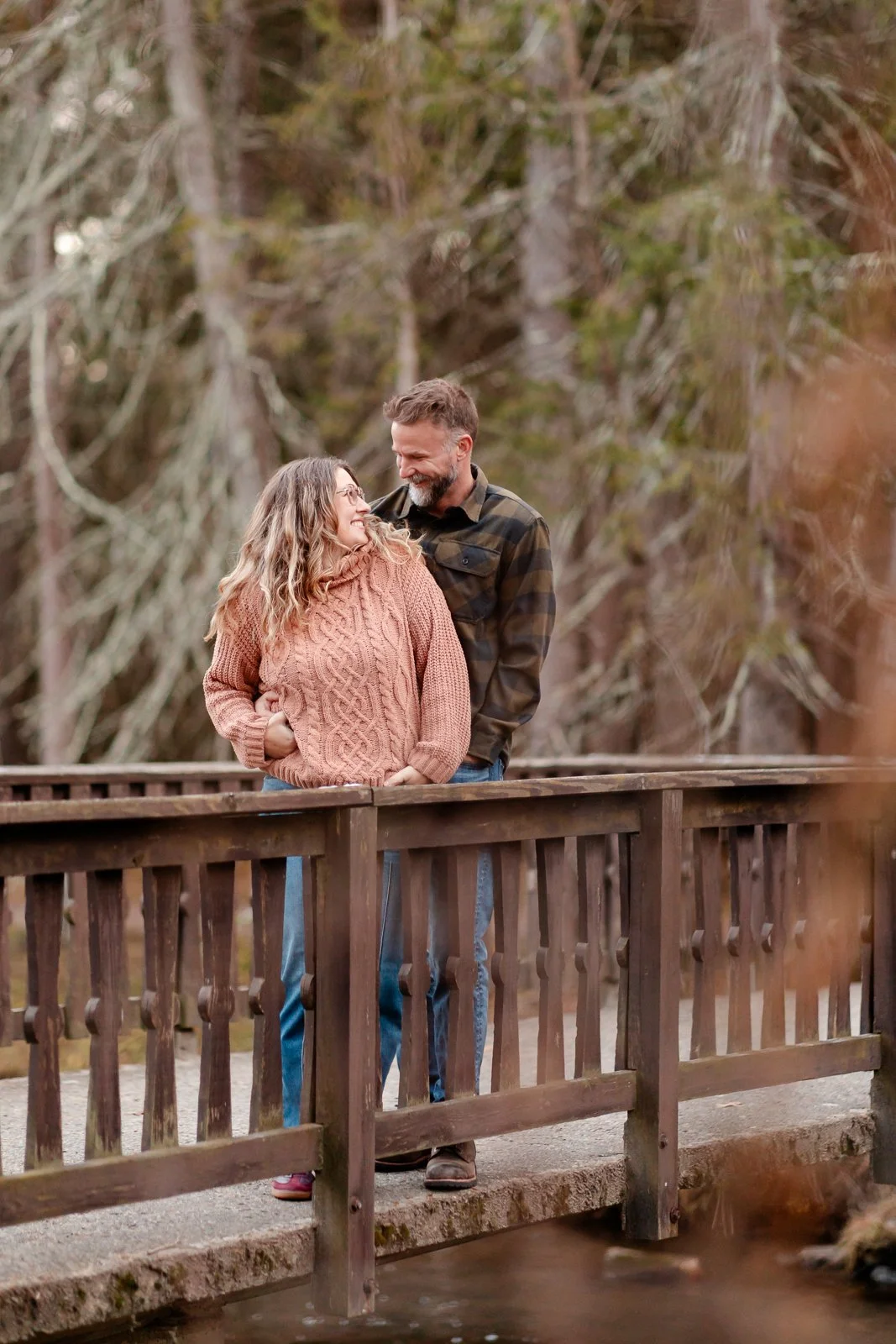 A couple standing on a wooden bridge in a forest, looking at each other and smiling warmly.