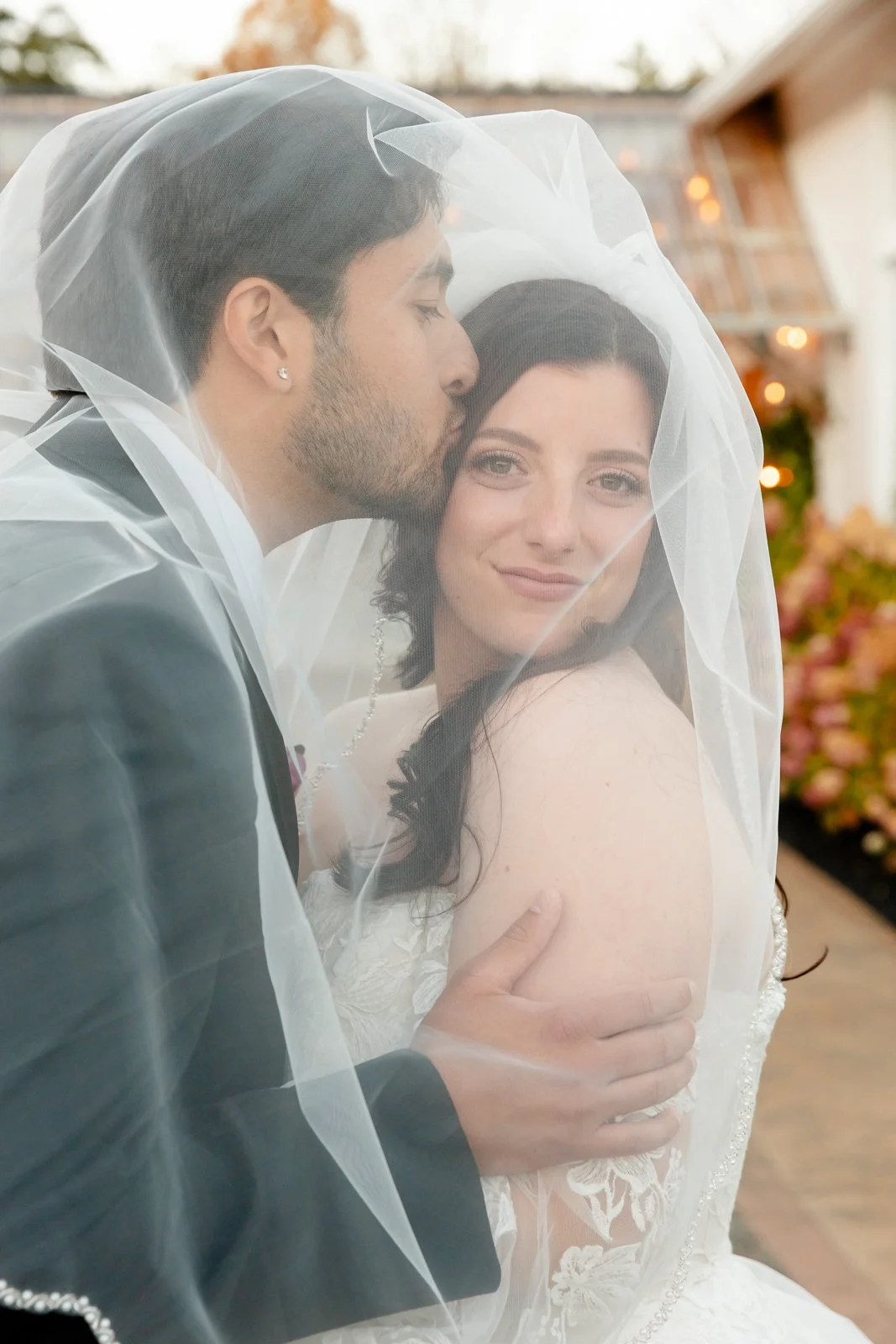 A newlywed couple embracing under a wedding veil, with the groom kissing the bride's forehead, outdoors with hanging lights and flowers in the background.