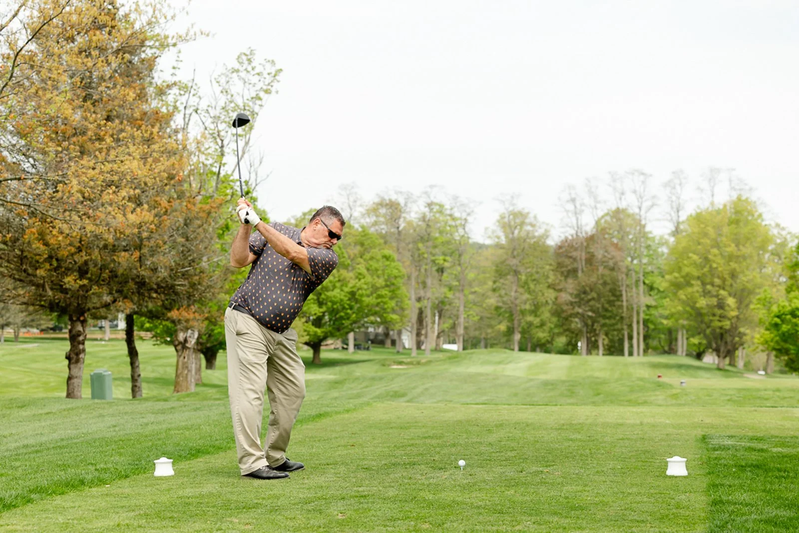 A man in outdoor clothing, wearing sunglasses and gloves, is swinging a golf club on a golf course surrounded by trees with green leaves.