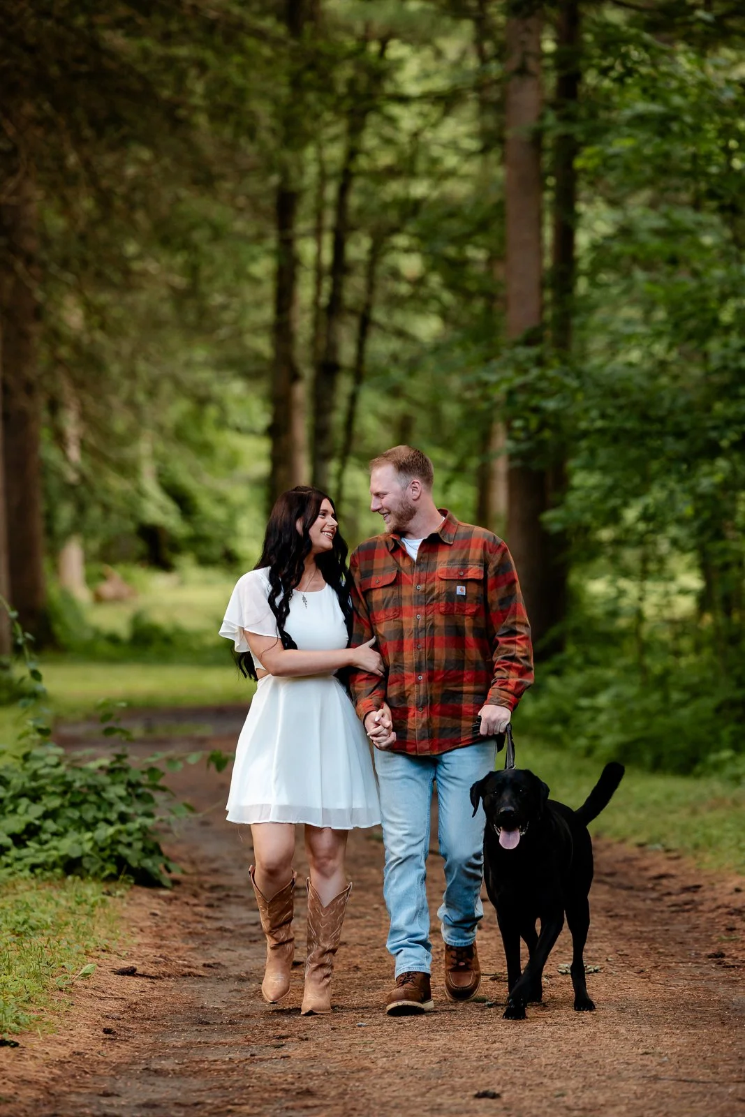 A couple walking with their black dog on a forest trail, smiling at each other, surrounded by green trees and foliage.