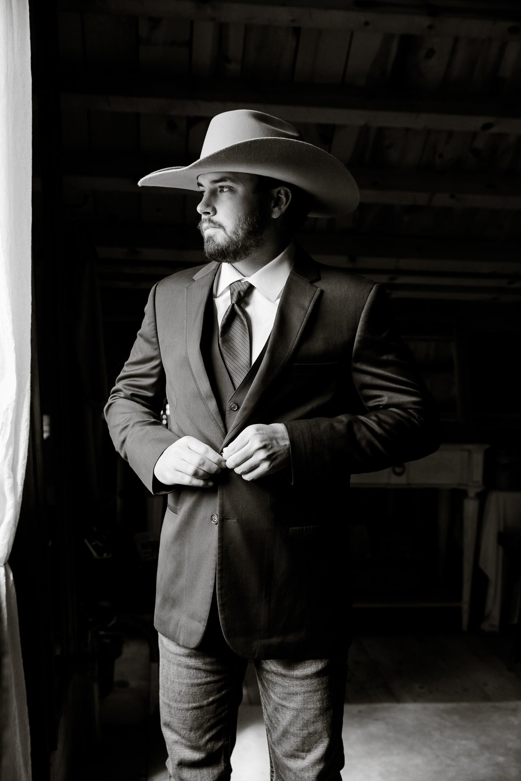 Black and white photo of a man in a suit and tie, wearing a cowboy hat, standing indoors near a window, gazing out with a contemplative expression.