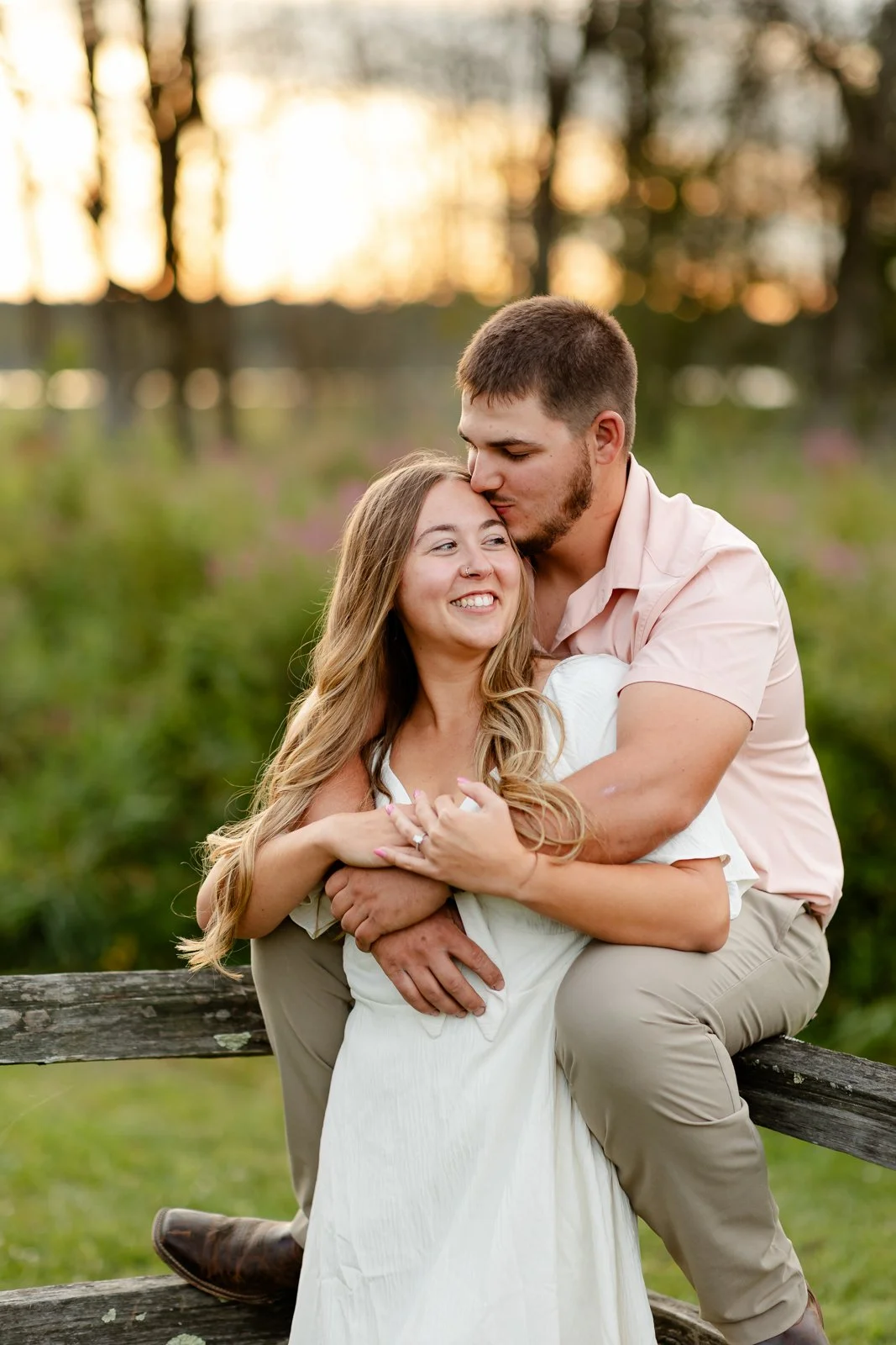A young couple sitting on a wooden fence in a park with trees and sunset in the background. The man is kissing the woman's forehead while hugging her from behind. The woman is smiling with her eyes closed, wearing a white dress, and the man is in a l