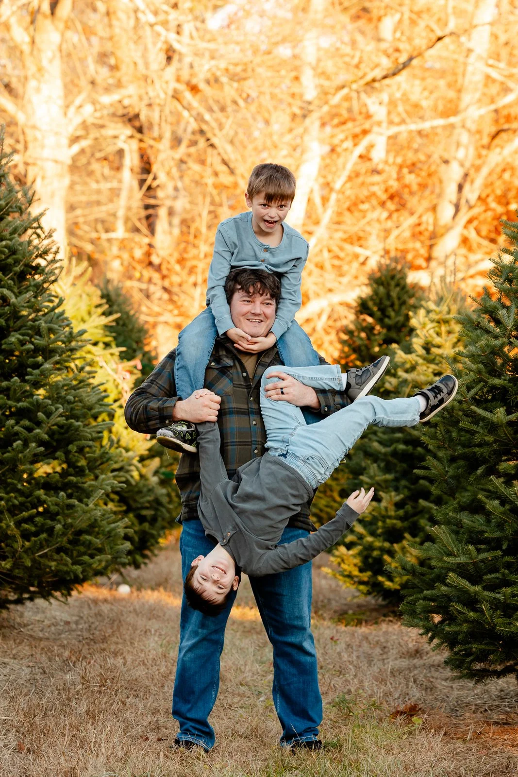 A man openly smiling while holding two boys, one on his shoulders and the other upside down, in a Christmas tree farm with autumn-colored trees in the background.