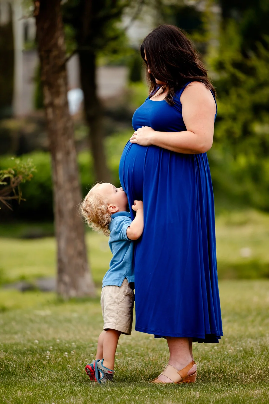 A pregnant woman in a blue dress is standing outdoors with a young boy hugging her belly and looking up at her.