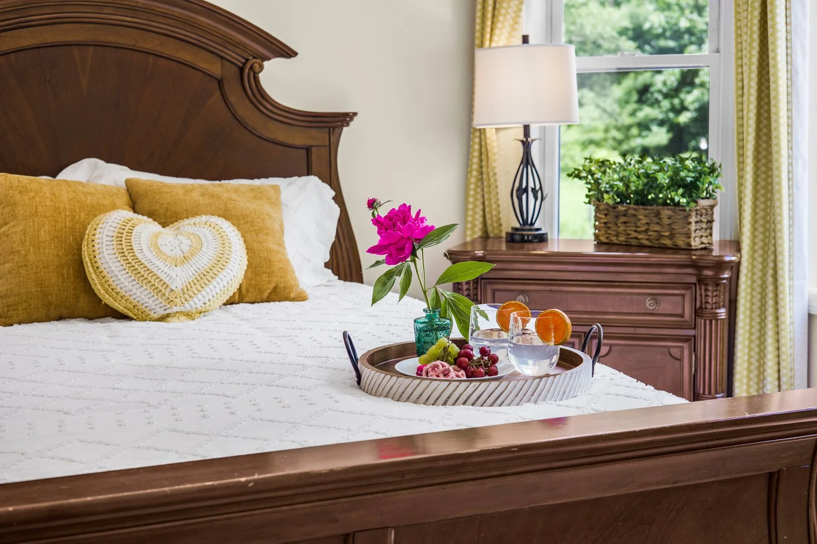 A bedroom with a wooden bed frame, white bedding, mustard yellow pillows, and a heart-shaped crocheted pillow. There is a tray on the bed with a pink flower in a green vase, two glasses of water with orange slices, and some fruit. A bedside table wit