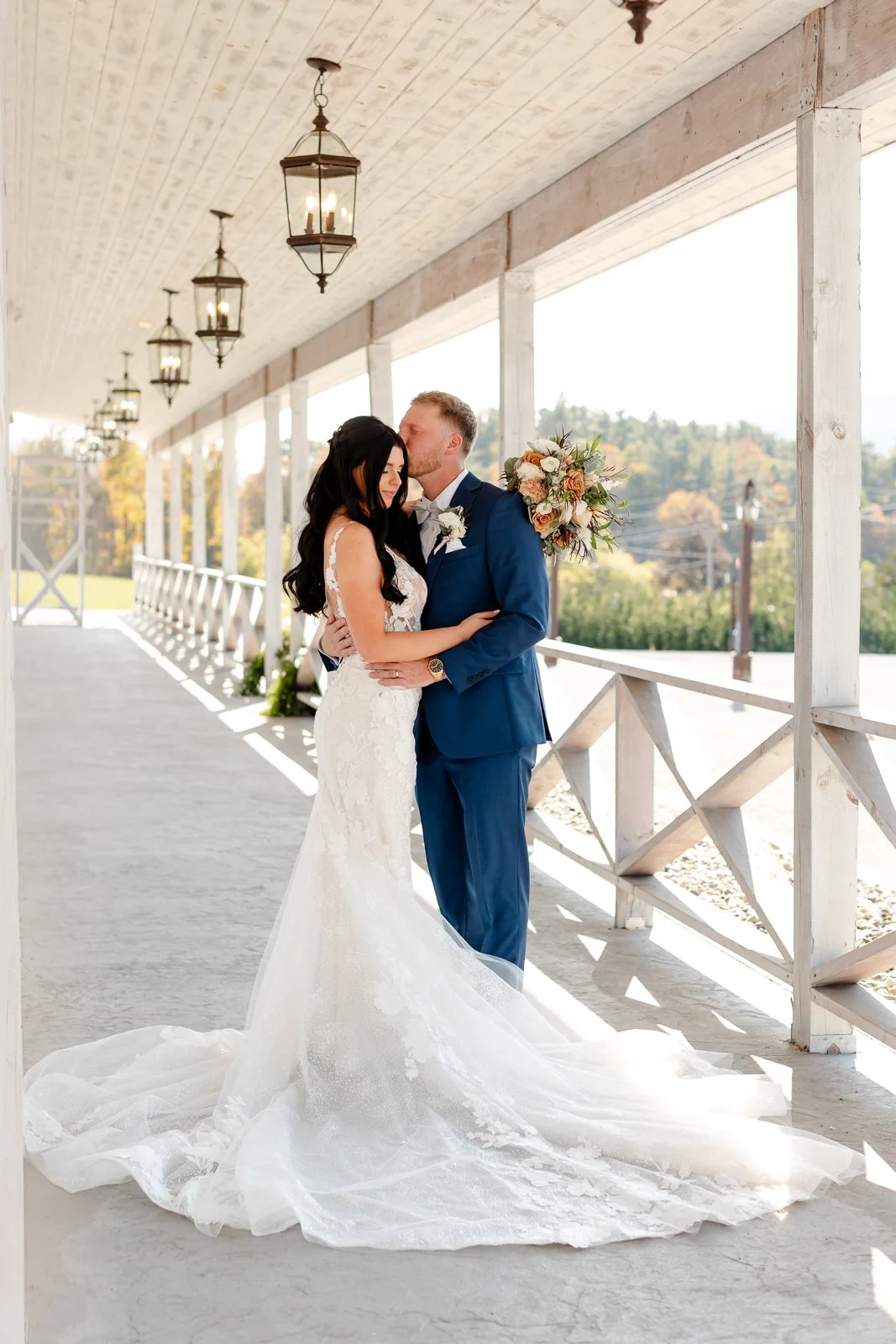 A bride and groom embracing on a porch during their wedding, with the groom kissing the bride on the forehead, holding a bouquet, and the bride wearing a white lace wedding dress.