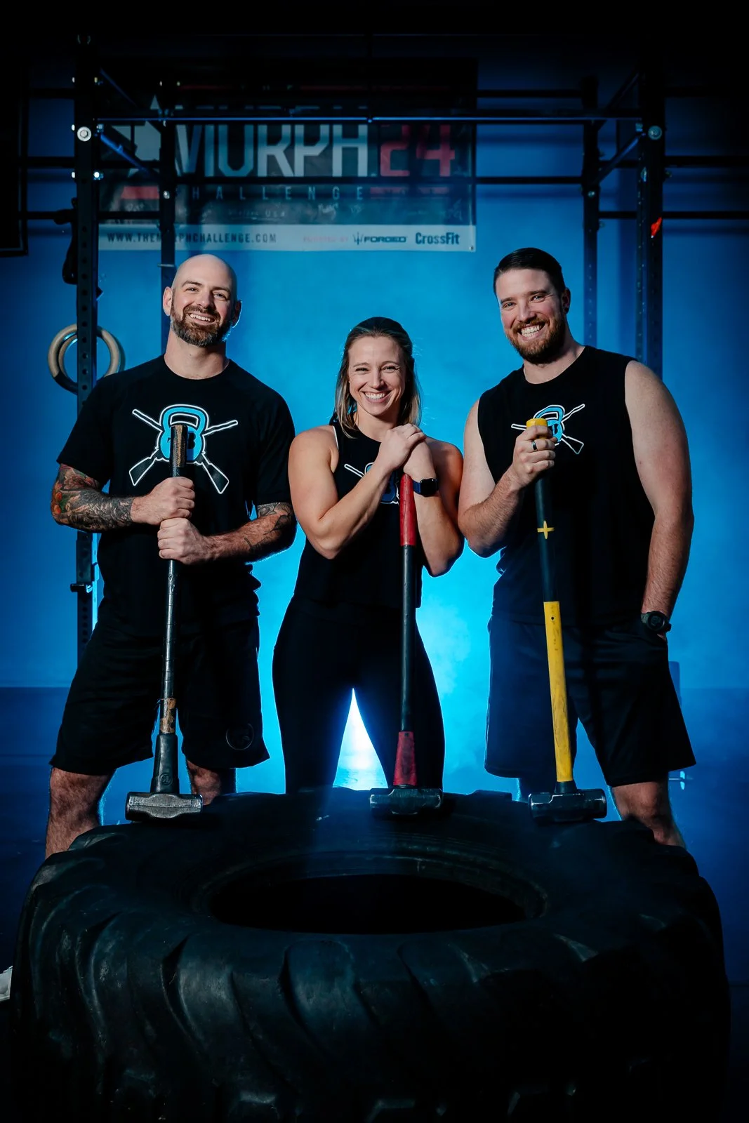 Three people in workout gear holding sledgehammers, standing in front of a large tire at a CrossFit gym, smiling at the camera with a blue background and a sign that reads 'VIKRH24 CHALLENGE'.
