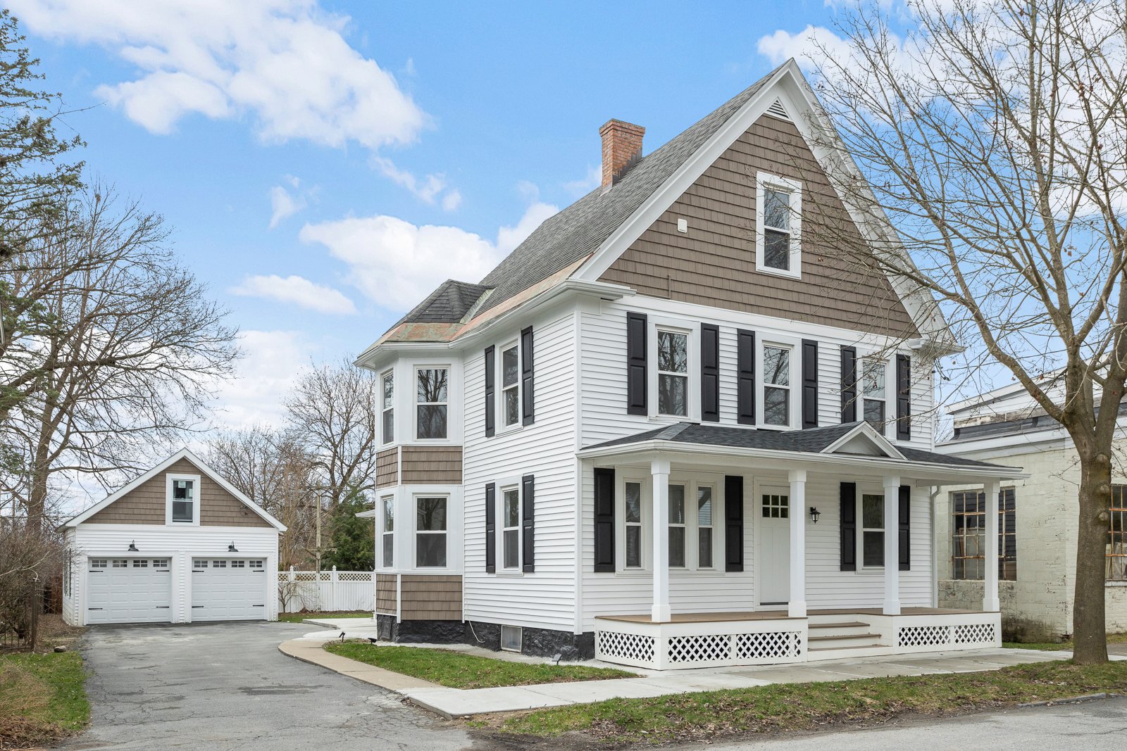 A large white and brown three-story house with a front porch, black shutters, and a detached garage in the backyard, on a suburban street with trees and a blue sky.
