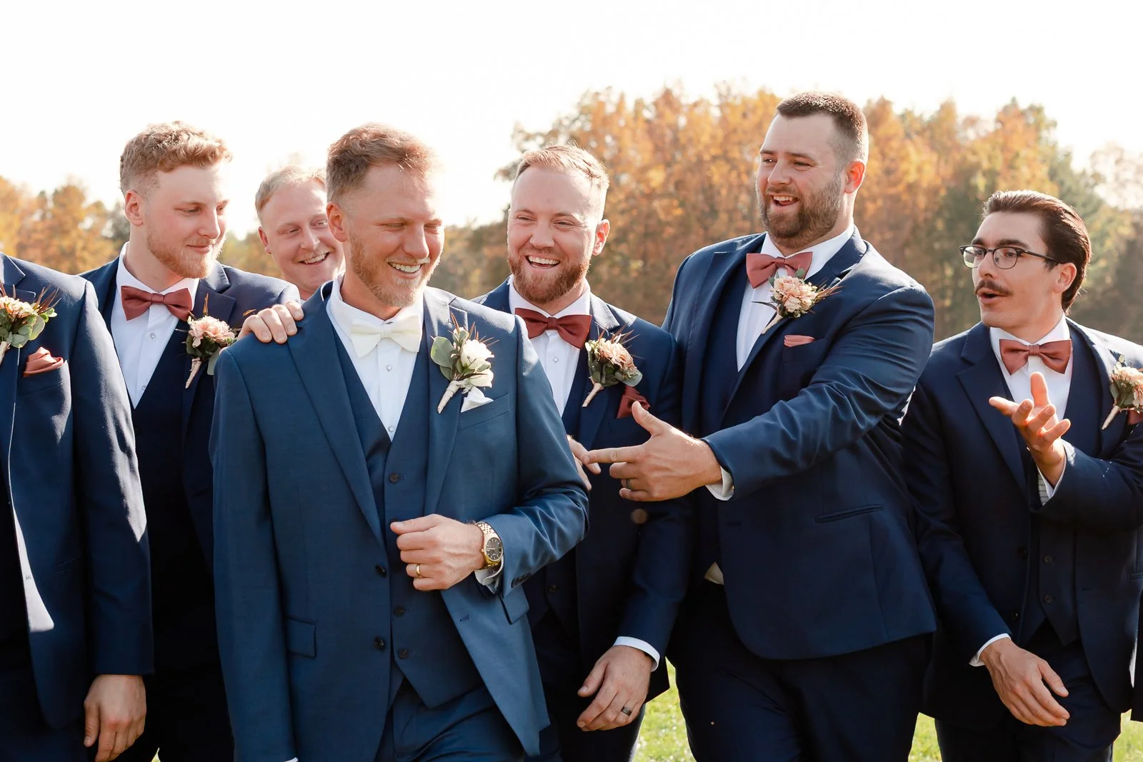 Group of men dressed in suits and bow ties celebrating outdoors with autumn trees in background.