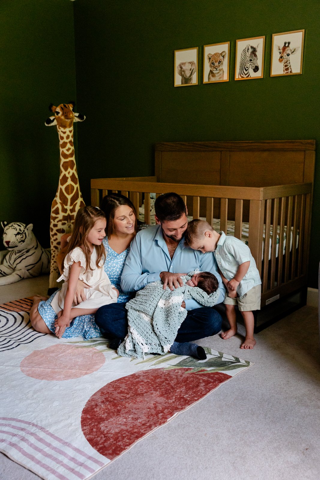 A family of five, including a man, woman, two children, and a baby, sitting on the floor in a nursery with green walls and animal-themed decor, celebrating the baby's arrival.