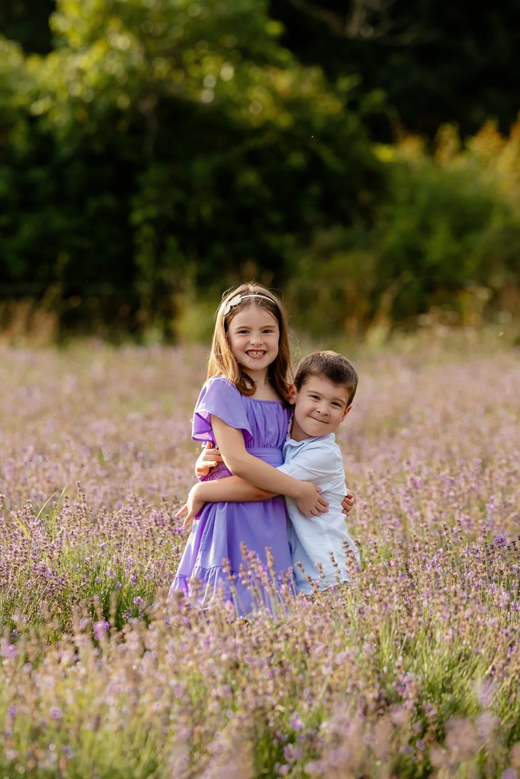 Two young children, a girl in a purple dress and a boy in a white shirt, hugging and smiling in a field of purple flowers with greenery in the background.