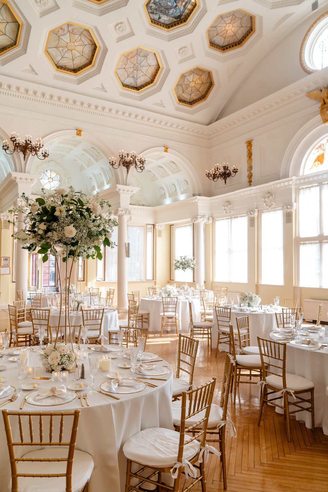 Elegant banquet hall decorated for a wedding reception with round tables covered in white tablecloths, gold chairs, floral centerpieces, and a high ceiling featuring decorative panels and chandeliers.