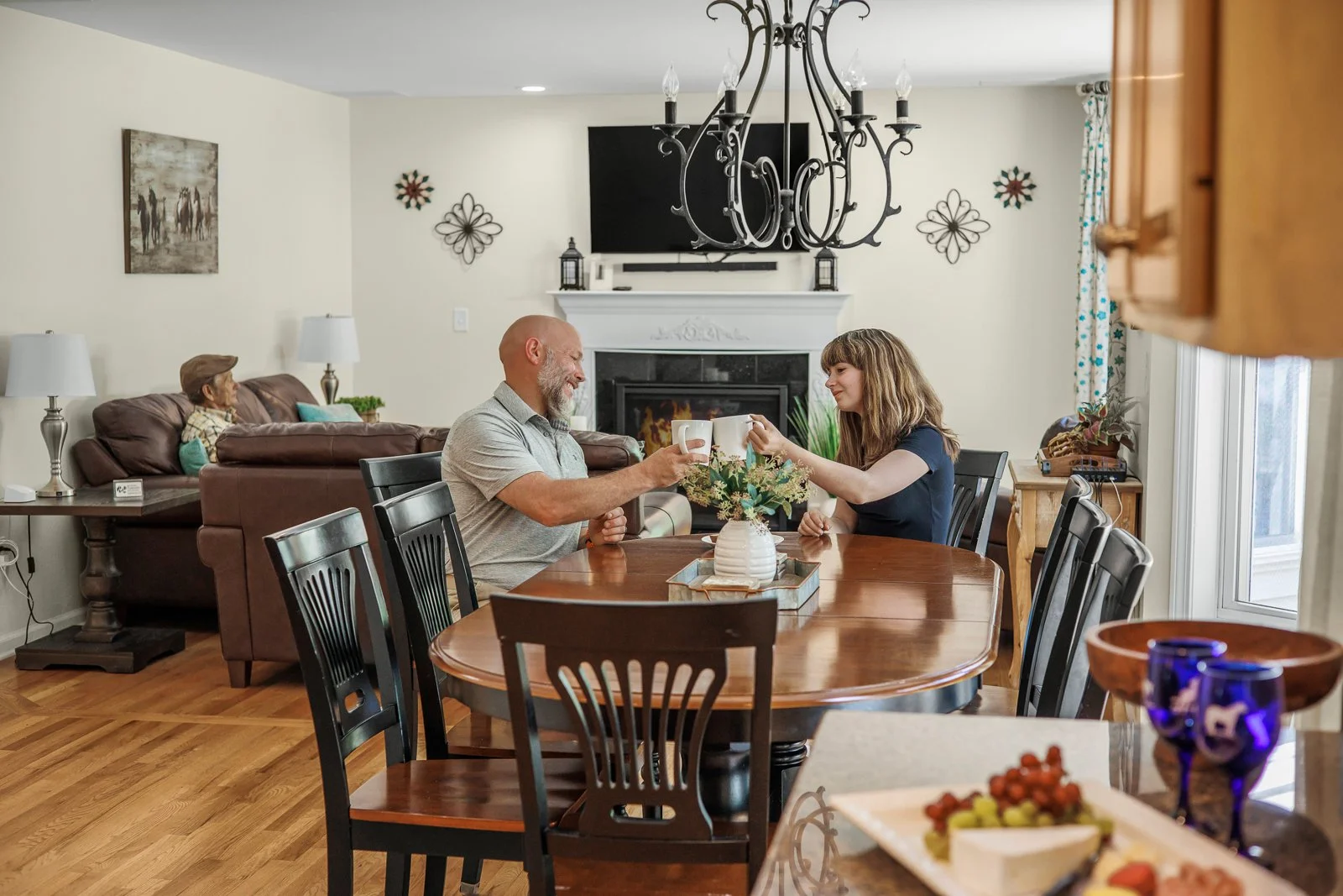 A family in a living room, with a man and woman sitting at a dining table clinking mugs, and a child on a couch in the background. The room has a fireplace, artwork, and decorative items.