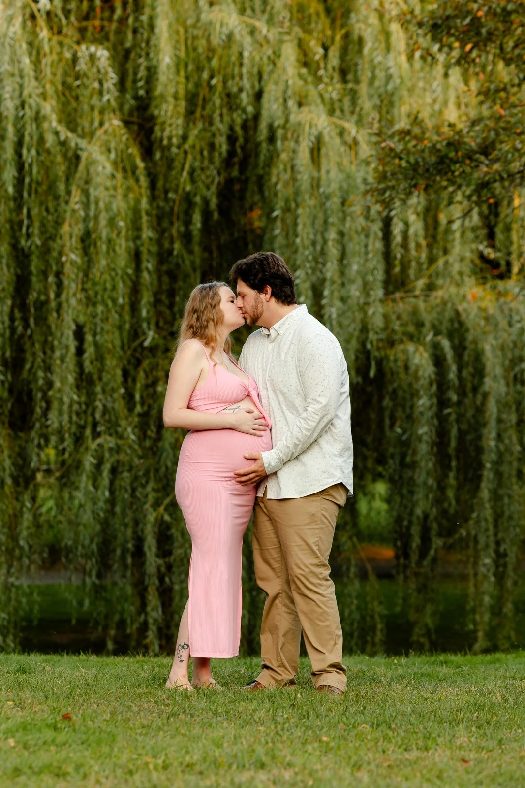 A pregnant woman and a man sharing a kiss outdoors, standing on grass with trees in the background.