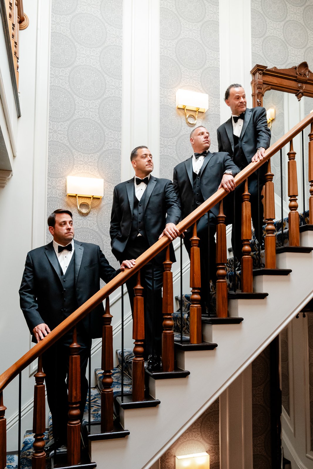 Five men dressed in black tuxedos with bow ties standing on a staircase in an elegant interior, looking in different directions.