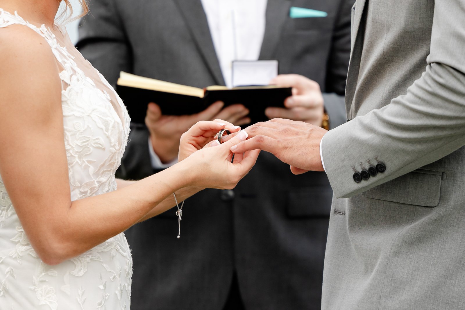 A bride and groom exchanging wedding rings during a wedding ceremony with an officiant in the background holding a book.