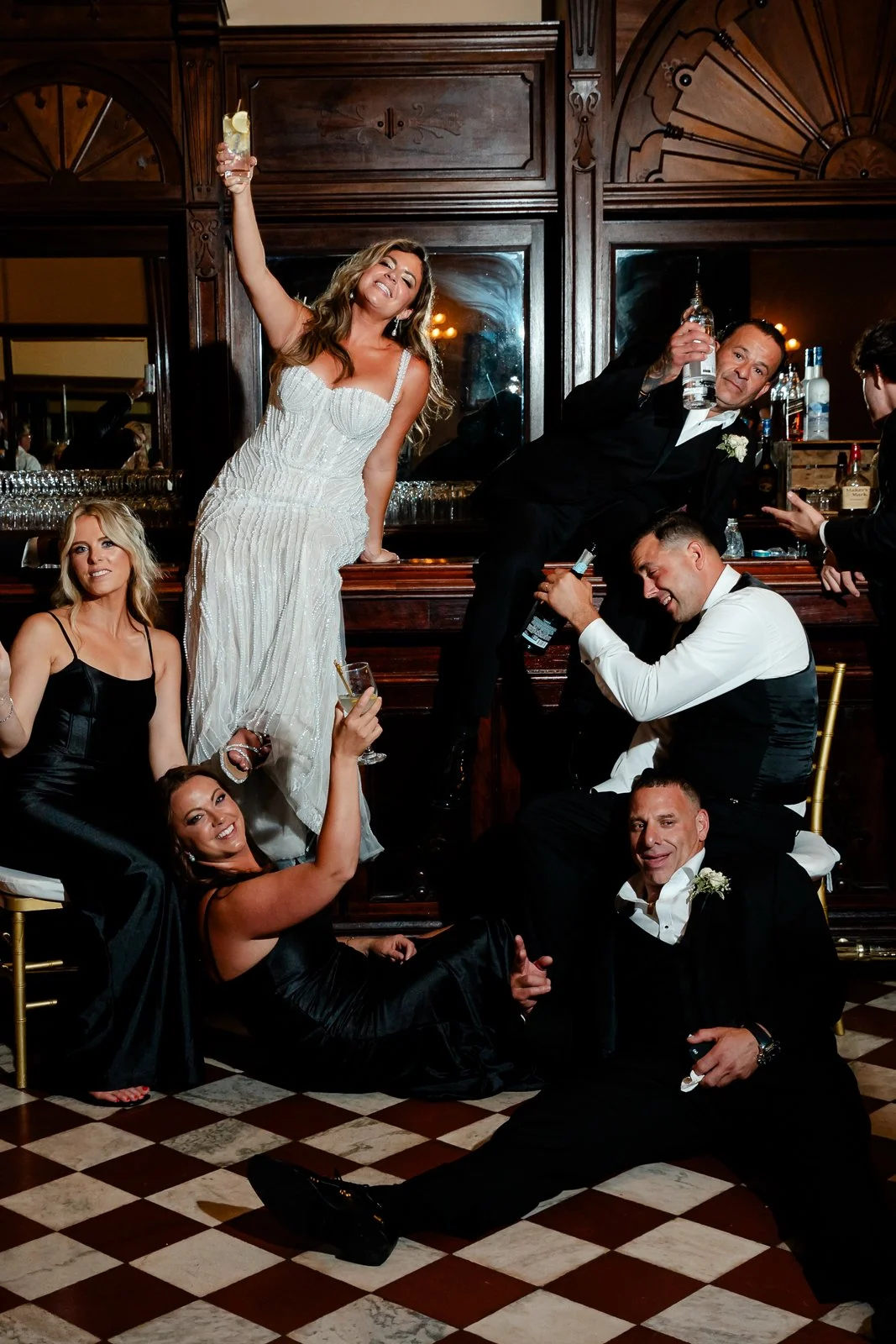 Group of people celebrating at a wedding reception, with some posing on the floor and others on the bar, holding drinks and making toasts.