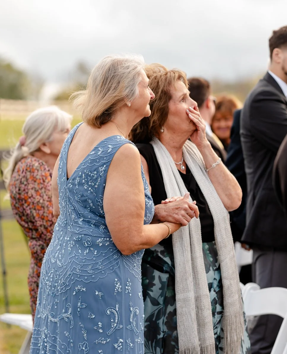 Two women, one in a blue embroidered dress and the other in a black top with a light scarf, appear emotional at an outdoor gathering, possibly a wedding.