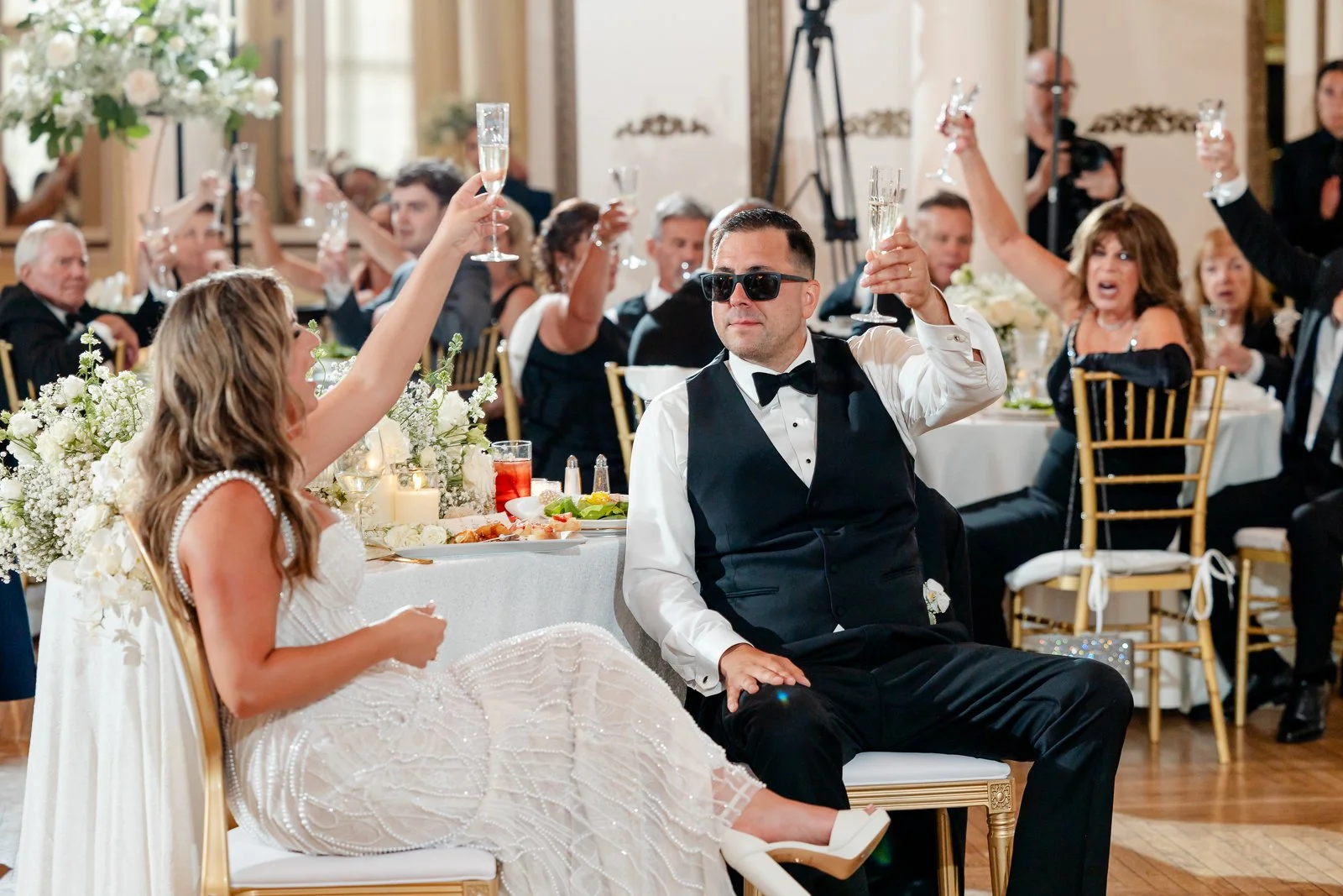 A wedding reception with a bride and groom raising champagne glasses, surrounded by guests at elegantly decorated tables.