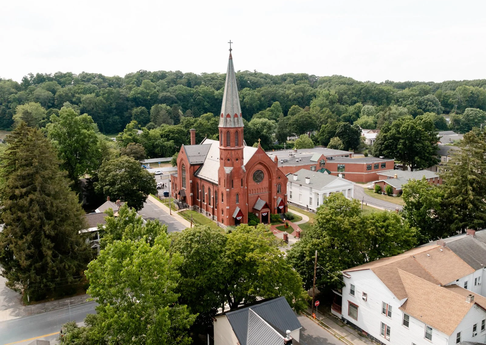 Aerial view of a red brick church with a tall steeple surrounded by trees and residential houses in a small town.