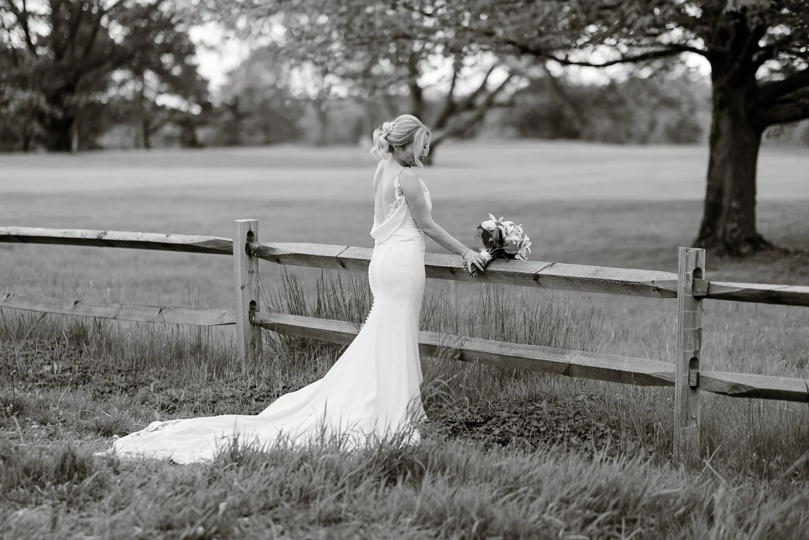 A bride in a white wedding dress standing alone by a wooden fence in an open grassy field with trees in the background, holding a bouquet of flowers resting on the fence.