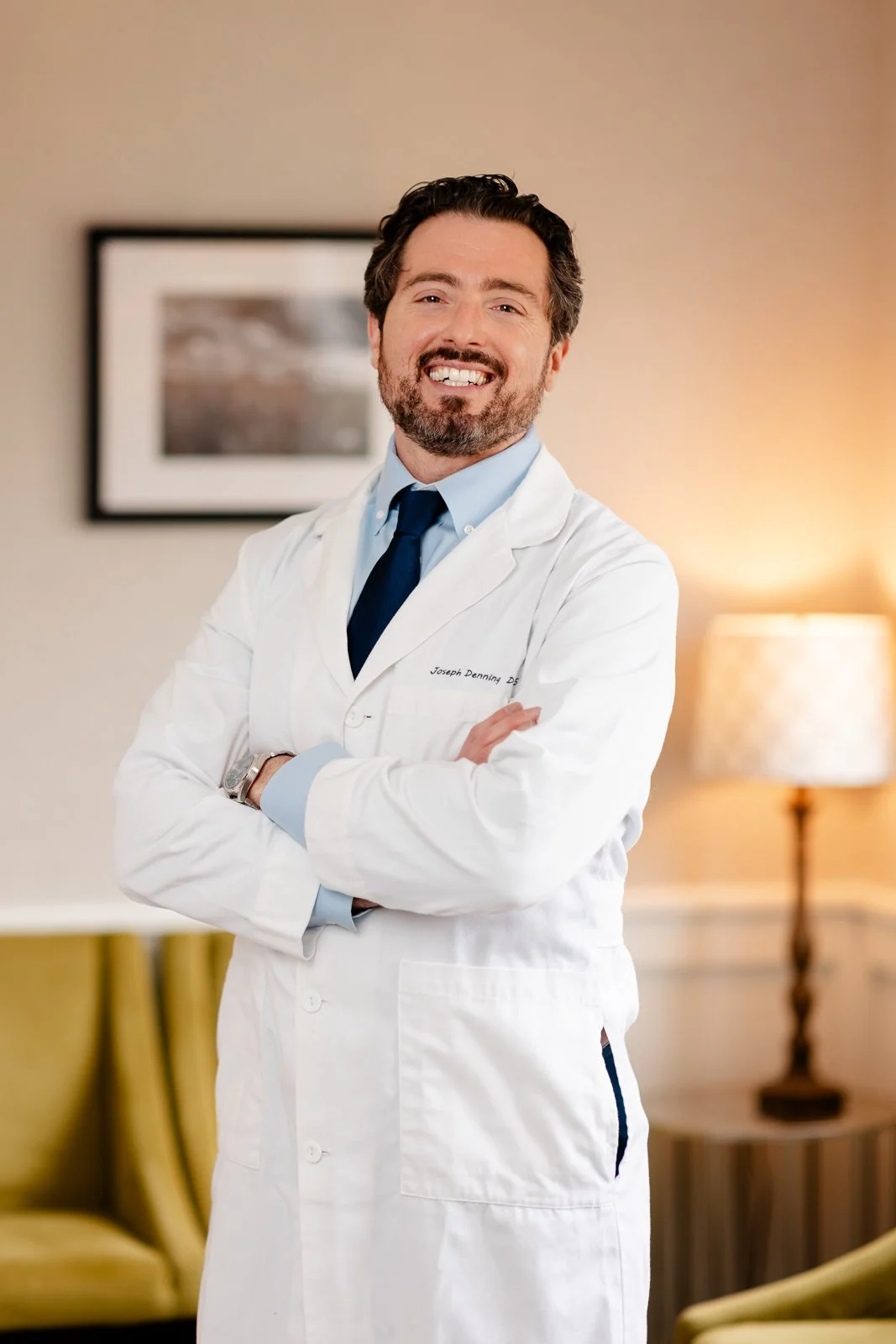 A smiling man with dark hair and beard, wearing a white medical coat with a name tag, light blue shirt, and dark blue tie, standing with arms crossed in a warmly lit room.