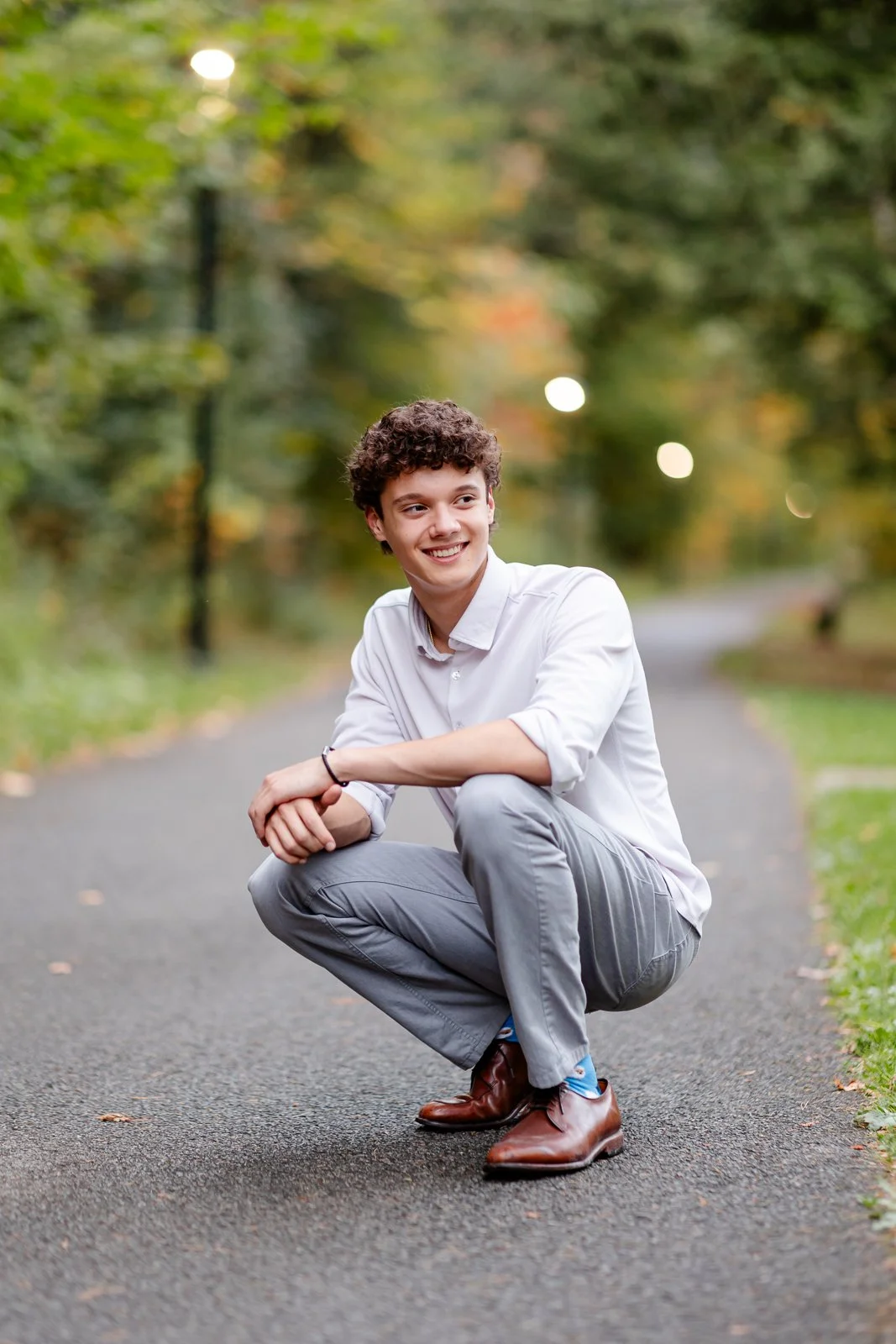 A young man with curly hair smiling and squatting on a paved path in a park with green trees and soft lighting.