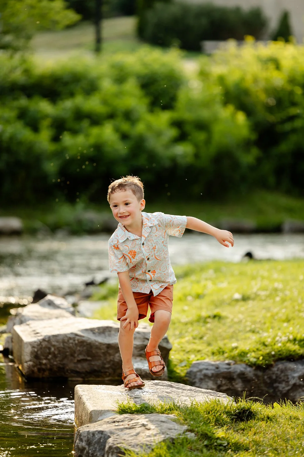 A young boy smiling and playing on stepping stones in a creek outdoors on a sunny day.