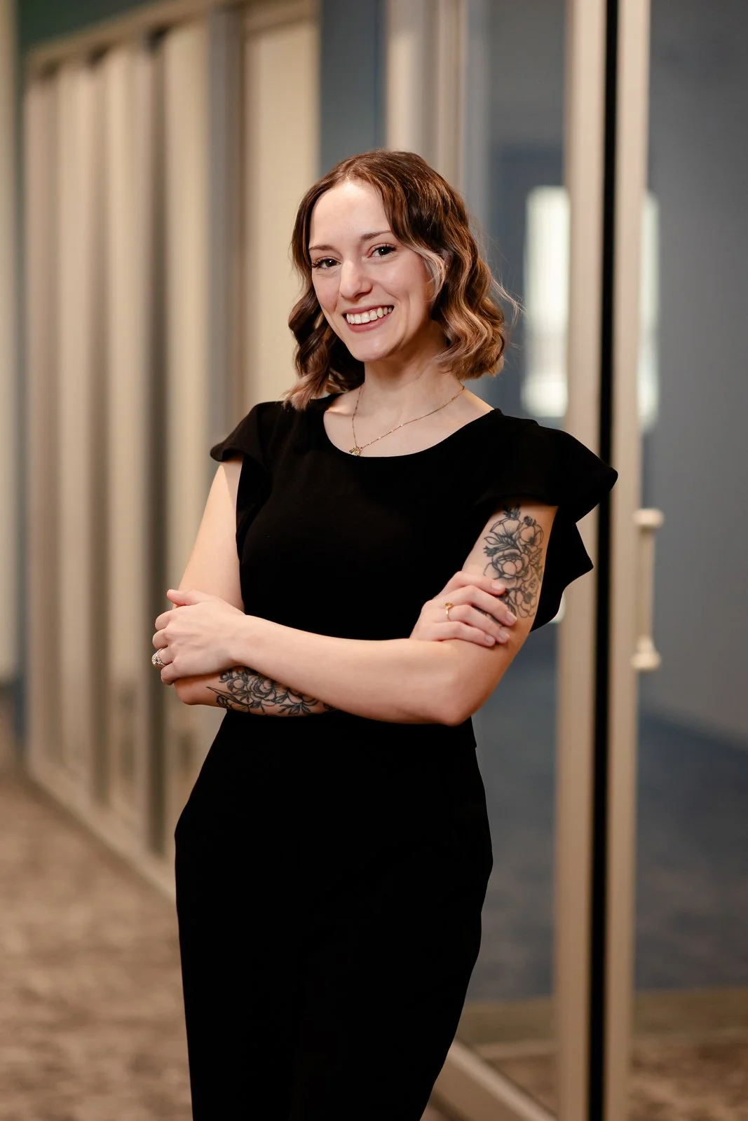 A young woman with shoulder-length wavy brown hair, wearing a black dress, standing in front of mirror doors, smiling with arms crossed, in an indoor setting.
