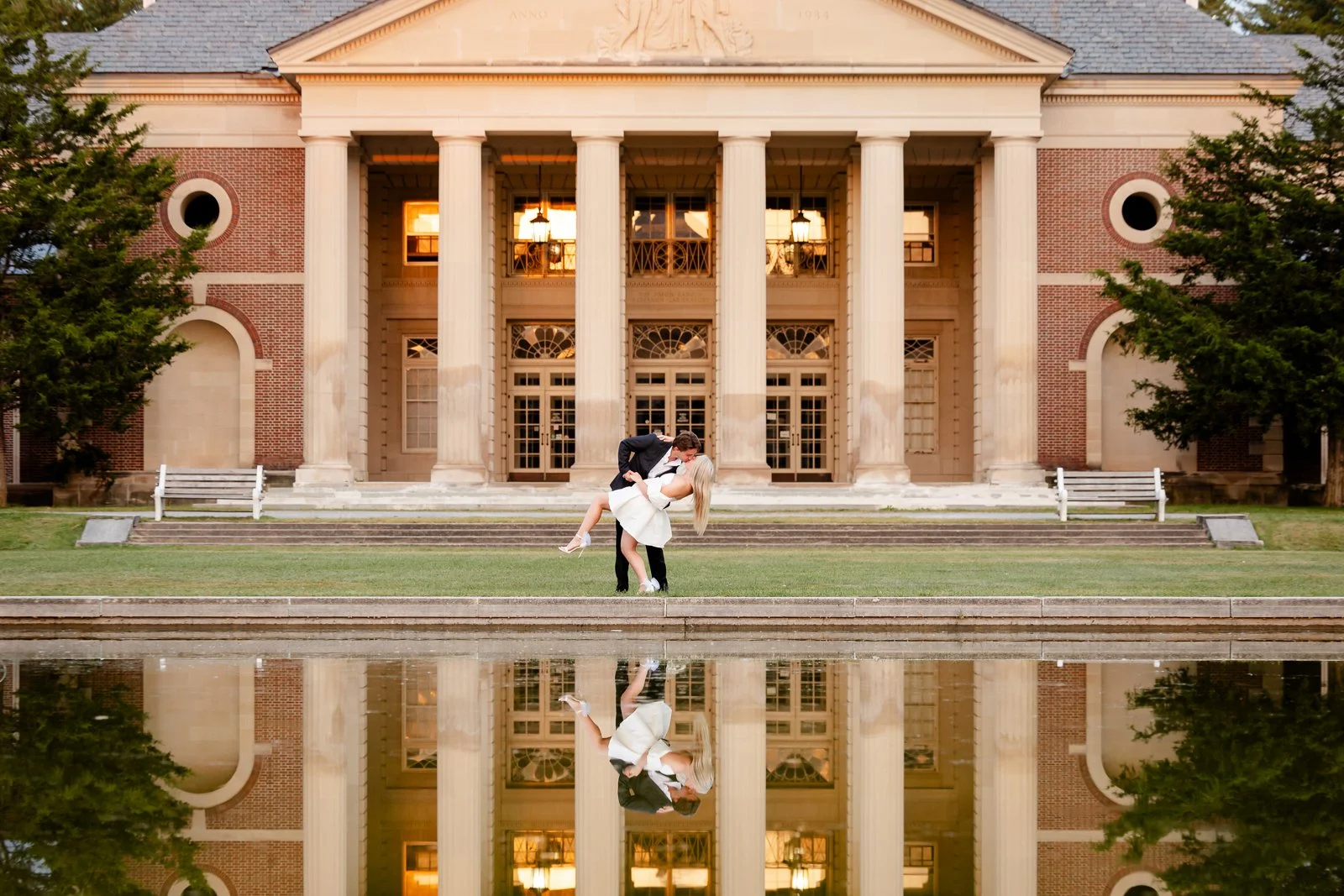 A couple dressed in wedding attire sharing a kiss and a dance in front of a large, historic building with tall columns, along a body of water that reflects the scene at sunset.
