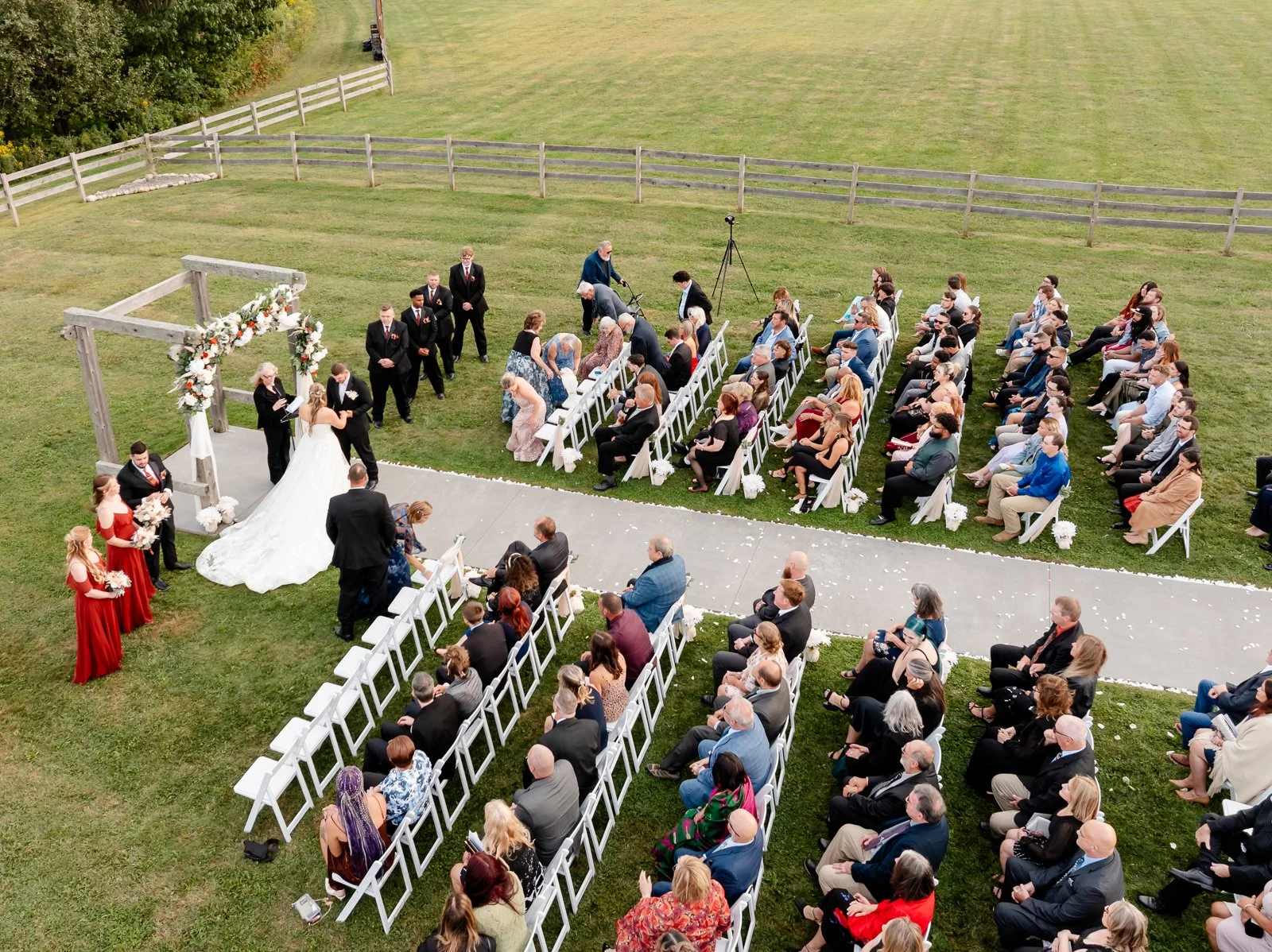 An outdoor wedding ceremony with a bride and groom standing under a wooden arch decorated with flowers, surrounded by guests seated in white chairs on a grassy field.