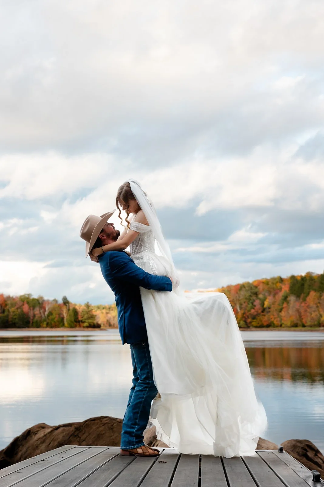 A man in a cowboy hat and blue jacket lifts a woman in a white wedding dress by a lake with autumn foliage in the background.