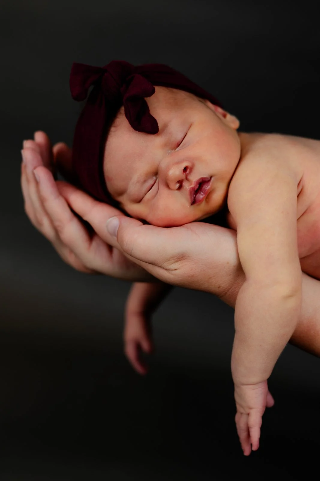 A newborn baby sleeping in an adult's hands with a dark background. The baby is wearing a maroon headband with a bow.