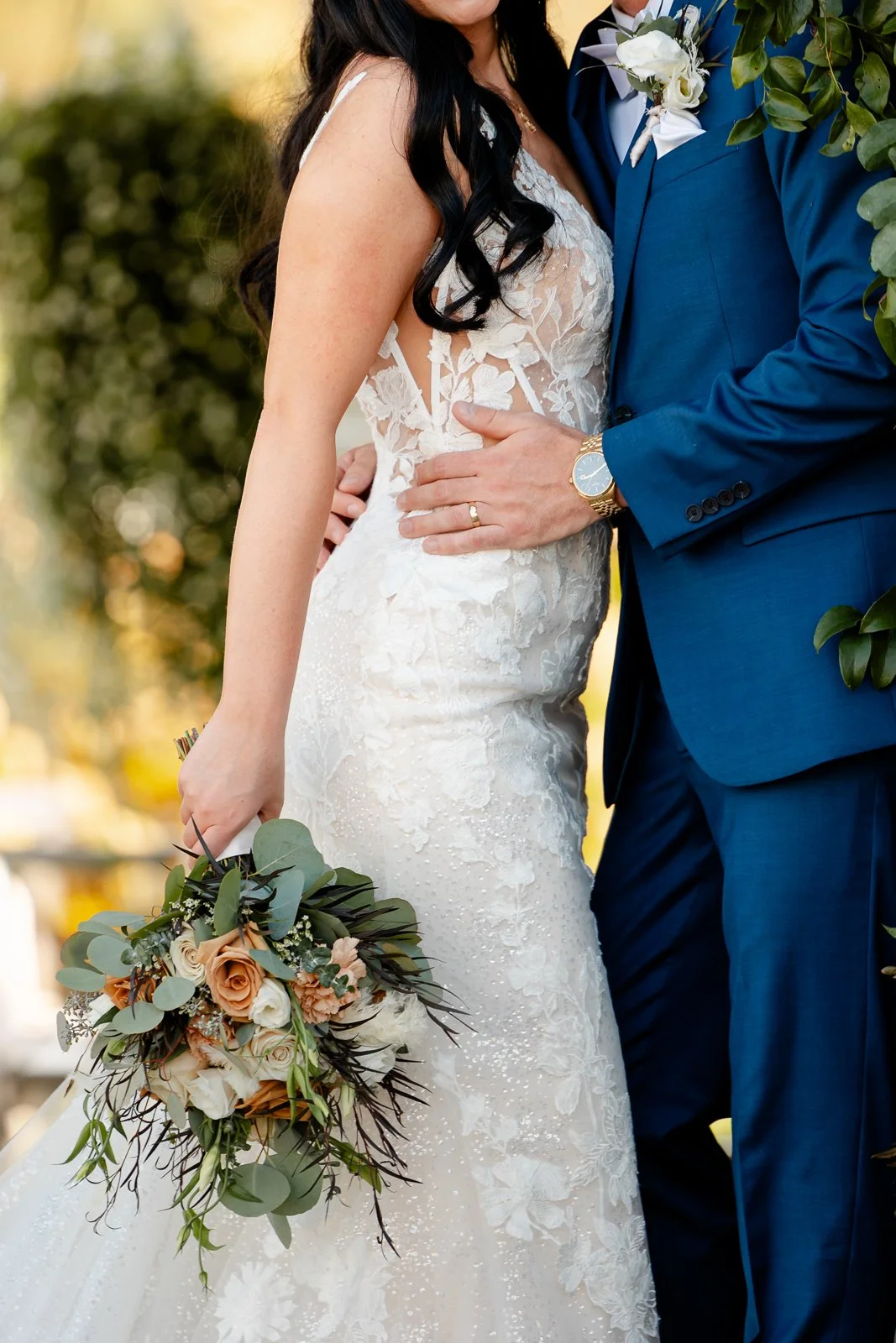 Close-up of a bride and groom during their wedding. The bride is in a white lace wedding dress holding a bouquet with roses and greenery. The groom is wearing a blue suit with a white boutonniere, and they are standing outdoors.