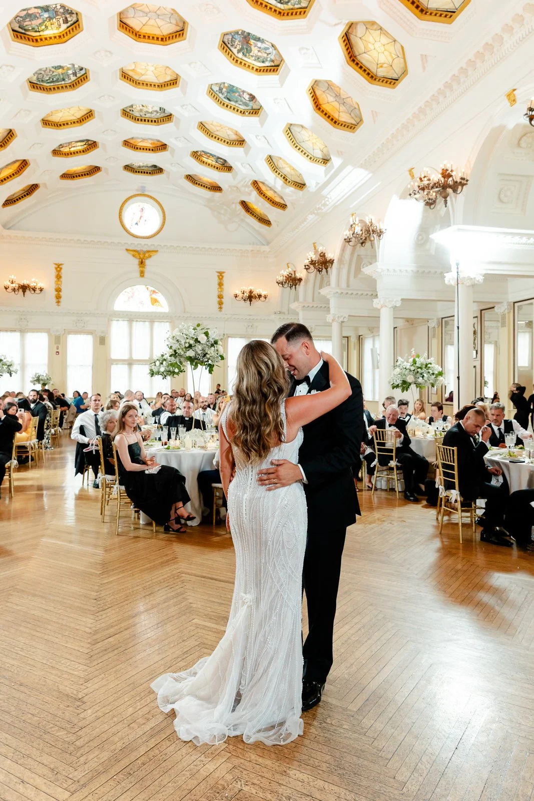 A bride and groom sharing a dance at their wedding reception in a grand, elegant hall filled with wedding guests seated at round tables.