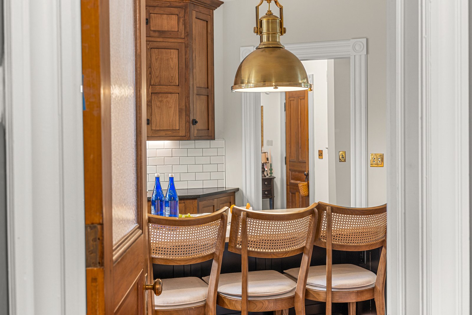 View of a cozy kitchen with wooden cabinets, white subway tile backsplash, three blue bottles on the counter, a gold pendant light hanging above a set of four wooden barstools, and a doorway leading to another room.