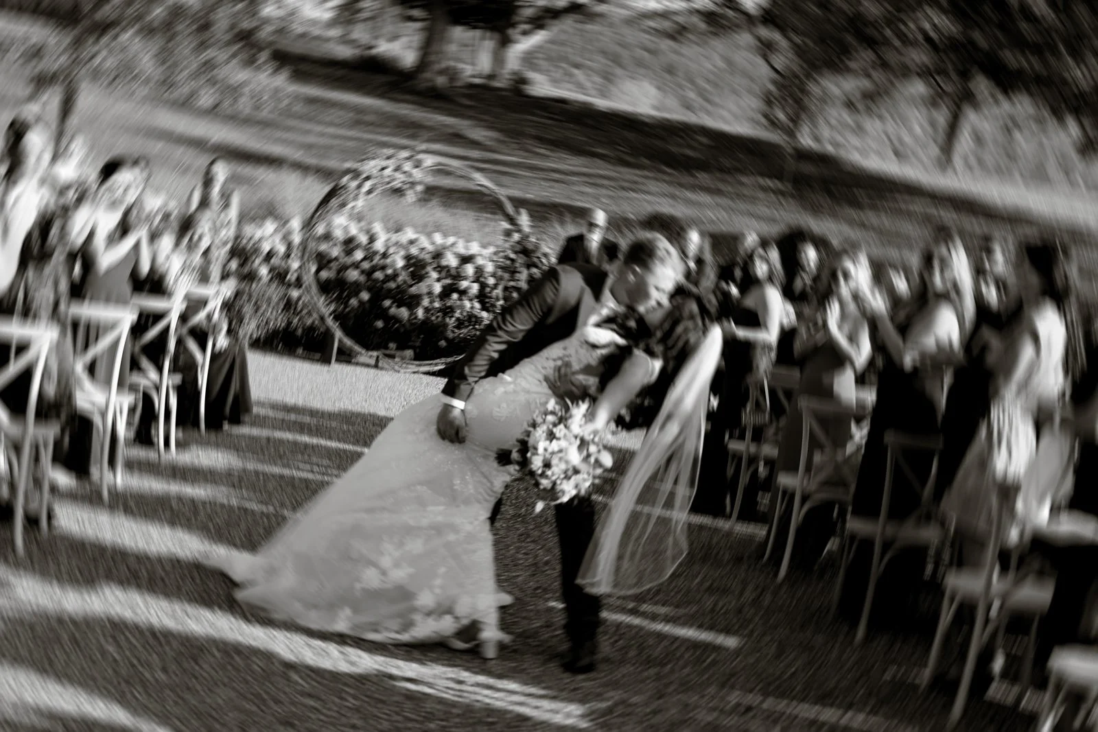 A bride and groom kiss at their outdoor wedding ceremony, surrounded by seated guests on either side of the aisle, with a decorative hoop and flowers in the background.