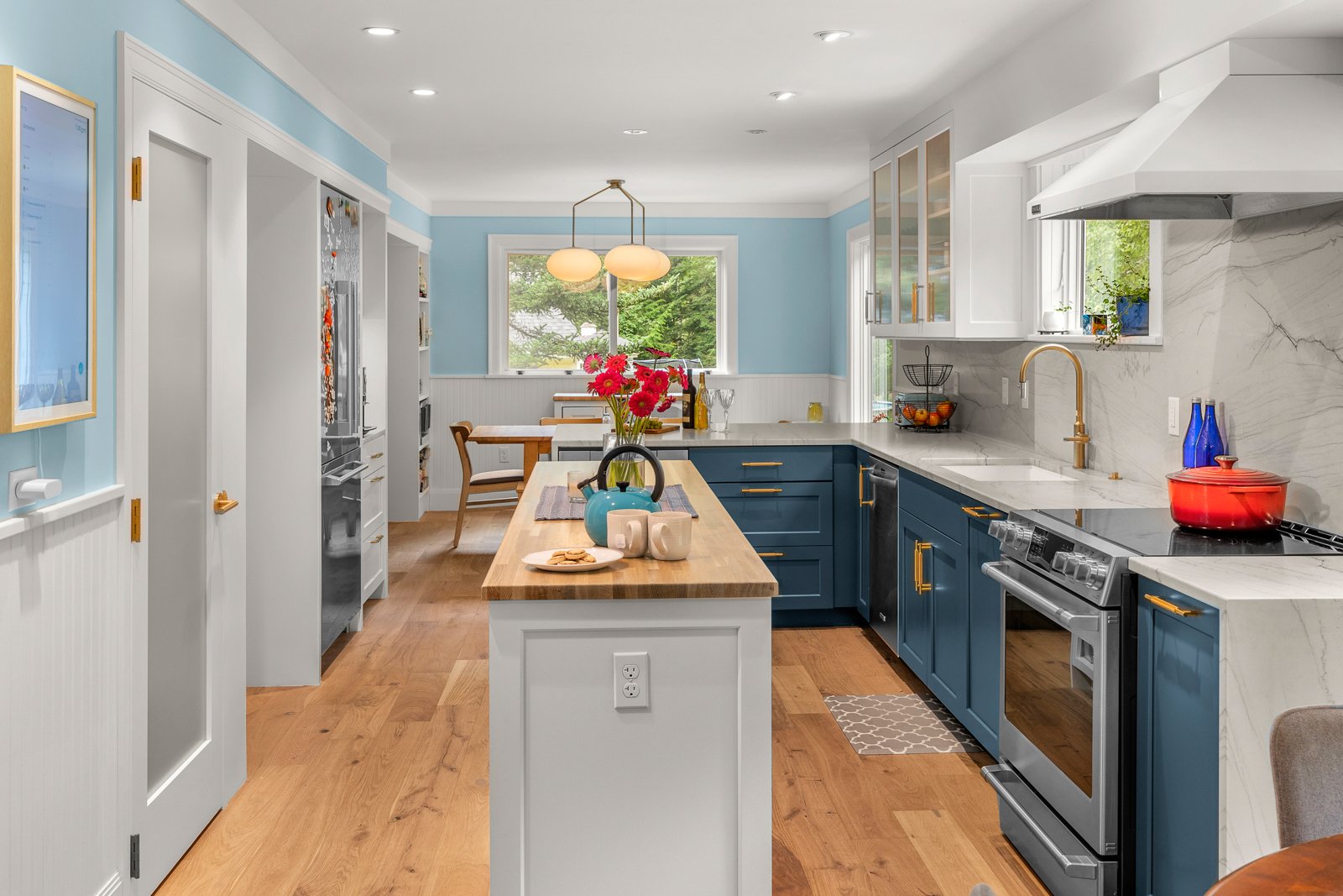 Kitchen with blue cabinets, white marble countertops, and a wooden dining table. There's a window with greenery outside, and a light fixture above the dining area. On the kitchen island, there is a blue teapot, flowers, mugs, and snacks. The room has