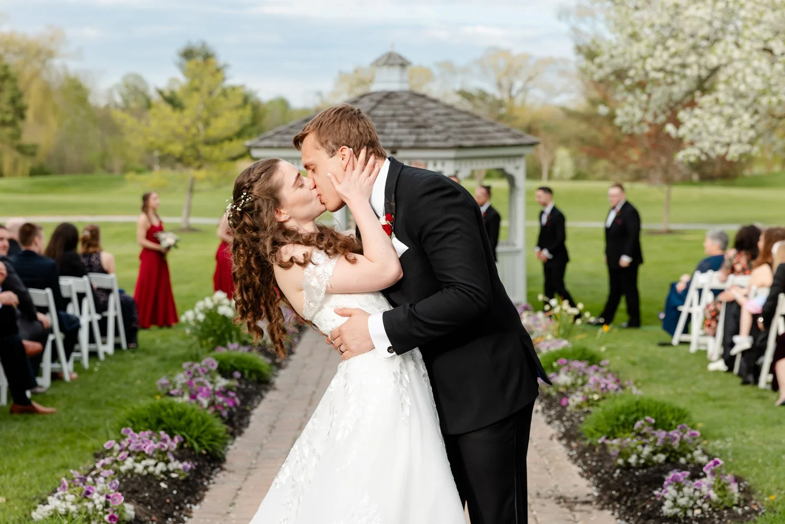 A newlywed couple sharing a kiss during their outdoor wedding ceremony, with guests seated on the sides and a gazebo in the background, on a spring day.