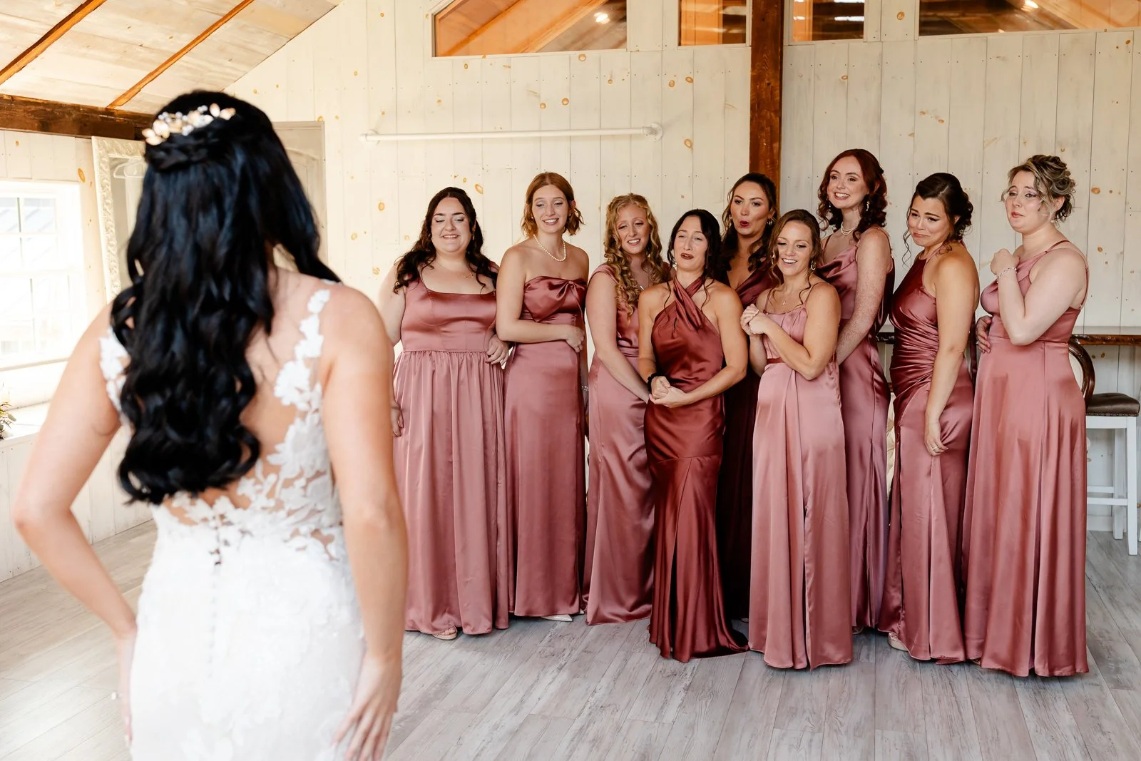 Bride and bridesmaids at a wedding preparing for the ceremony. The bride, with long black hair and a white lace dress, faces a group of ten women in matching rose-colored dresses inside a wooden venue.