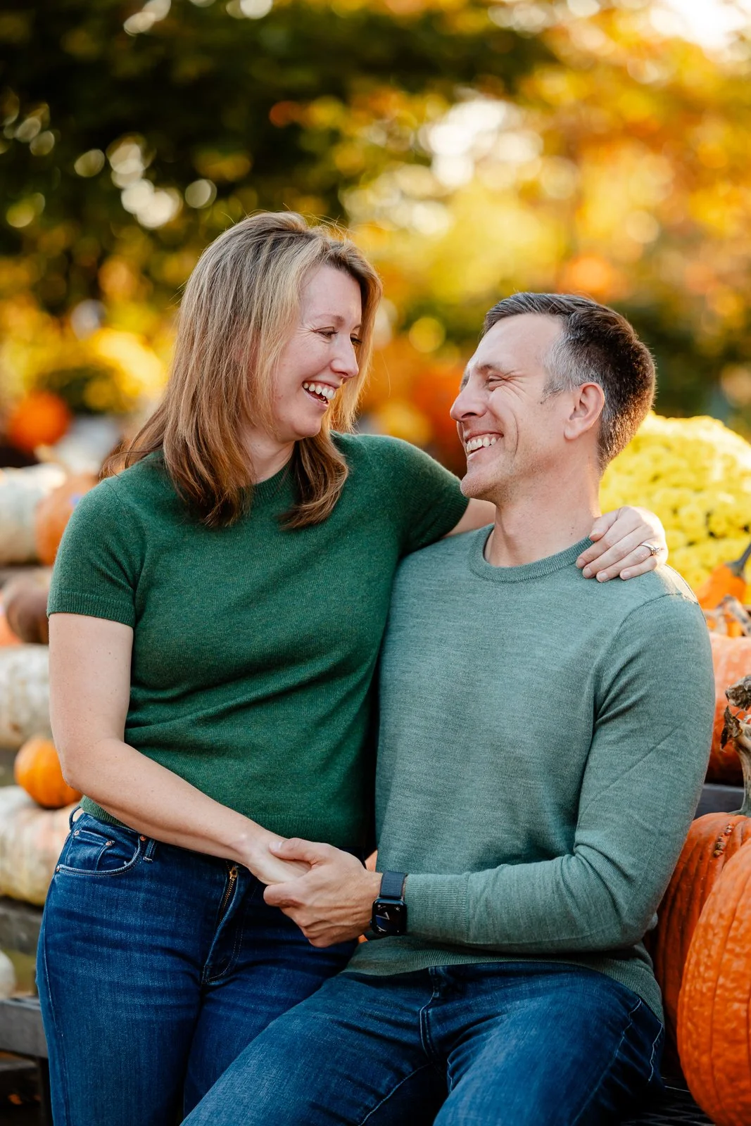 A happy couple laughing and holding hands at a pumpkin patch with autumn decorations and pumpkins in the background.